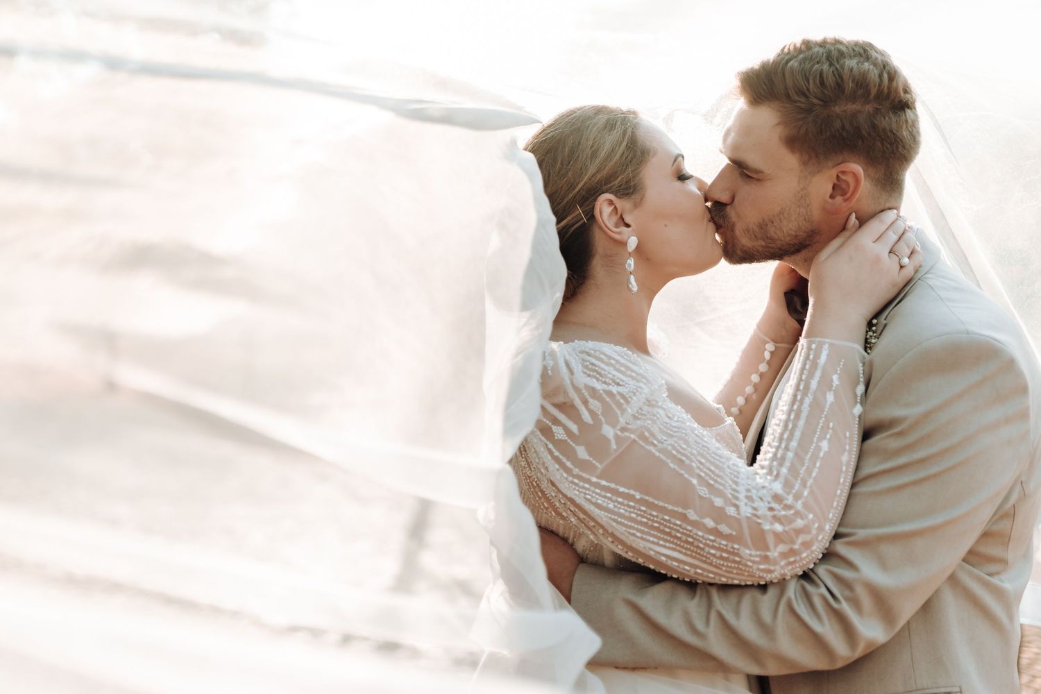 Romantic wedding kiss captured in soft sunlight with bride's lace sleeves and flowing veil.