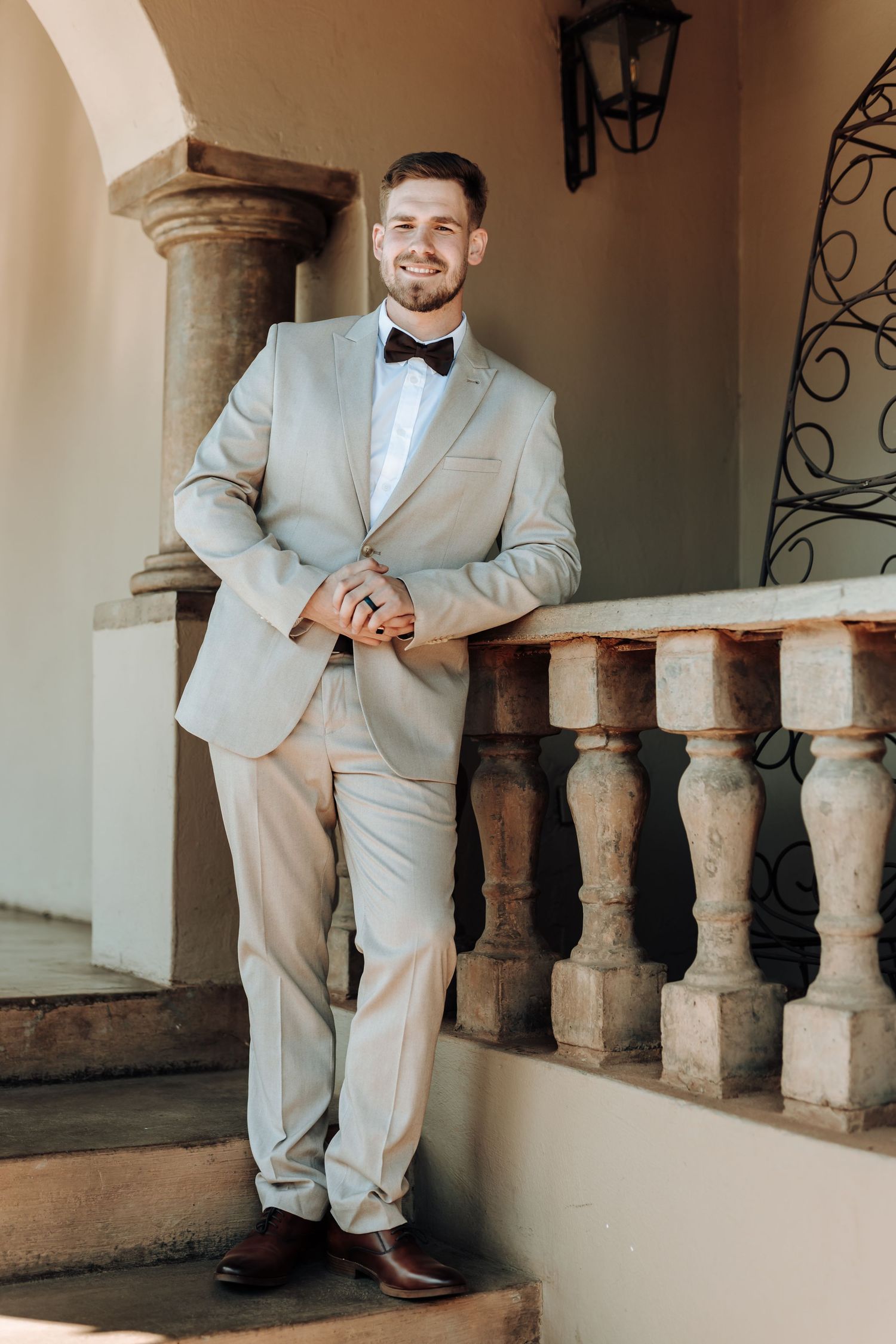 Someone in a light gray suit and bowtie poses on stone steps near ornate architecture.