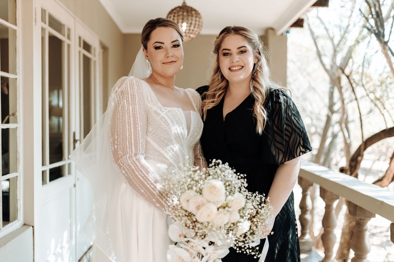 A bride in white lace gown poses with bridesmaid holding white floral bouquets.