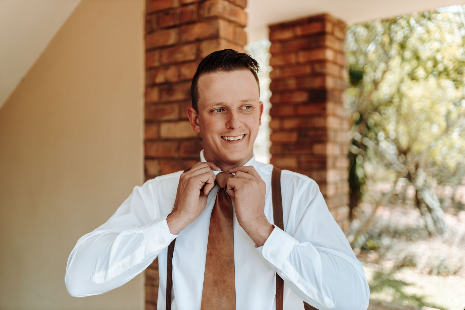 Someone adjusting a brown tie while wearing white shirt and suspenders.
