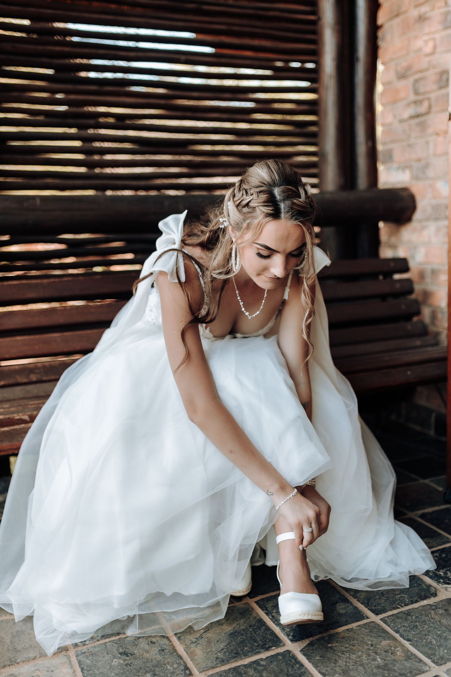 Bride in flowing white dress adjusts her shoe in rustic venue.