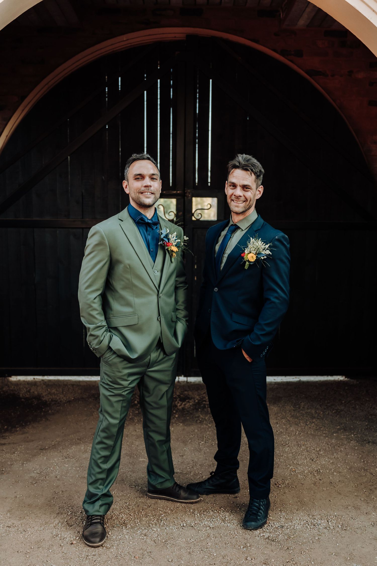 Two groomsmen in contrasting sage green and navy suits pose in front of a dark wooden doorway.