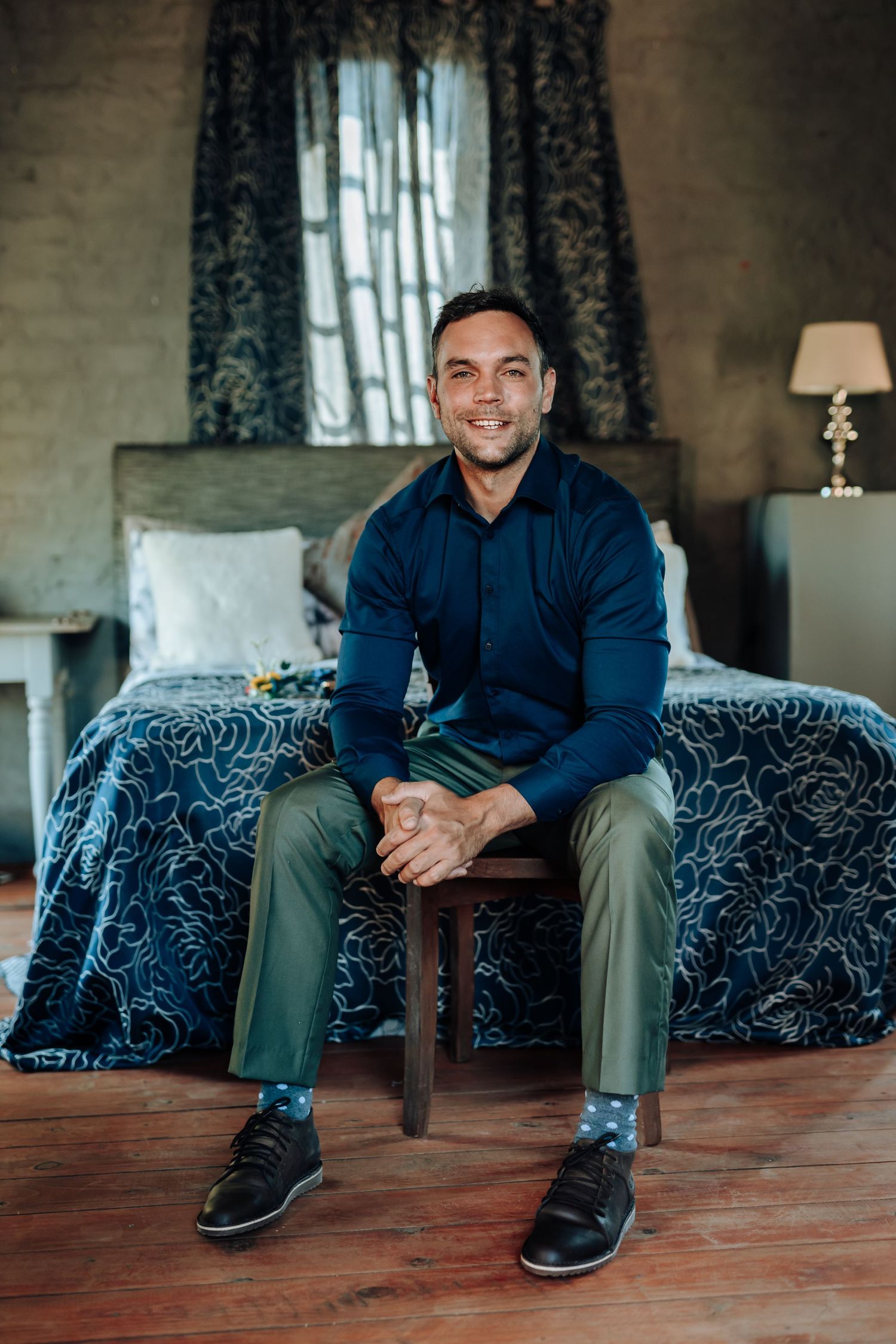 Well-dressed person seated on stool in bedroom with blue patterned bedding.