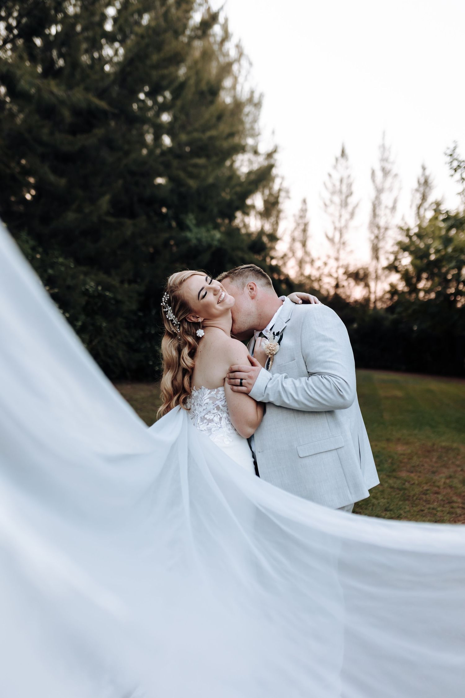 Dramatic wedding photo with flowing white dress against sunset and pine trees.