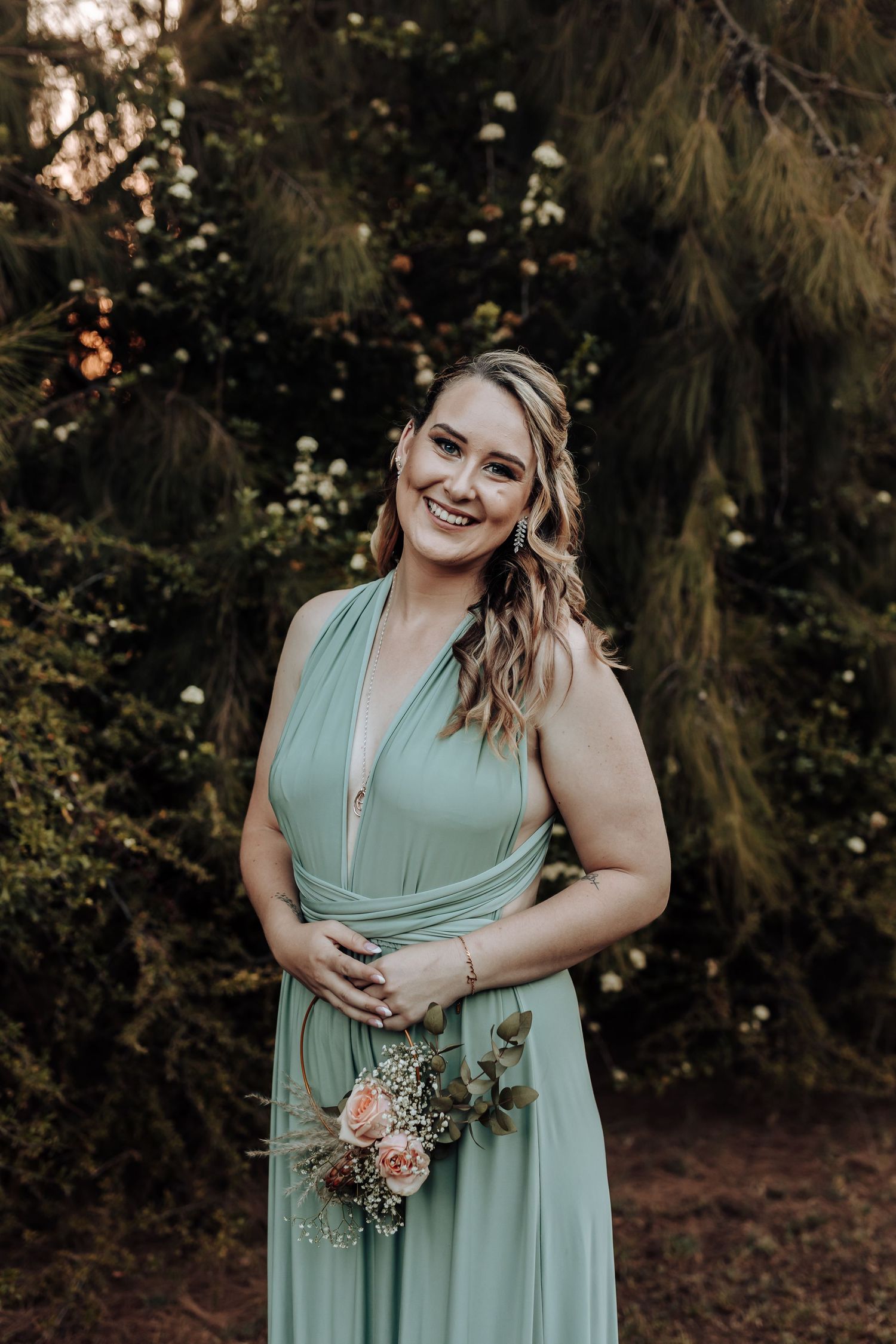 Bridesmaid in sage green wrap dress holds small floral bouquet while posing in garden setting.