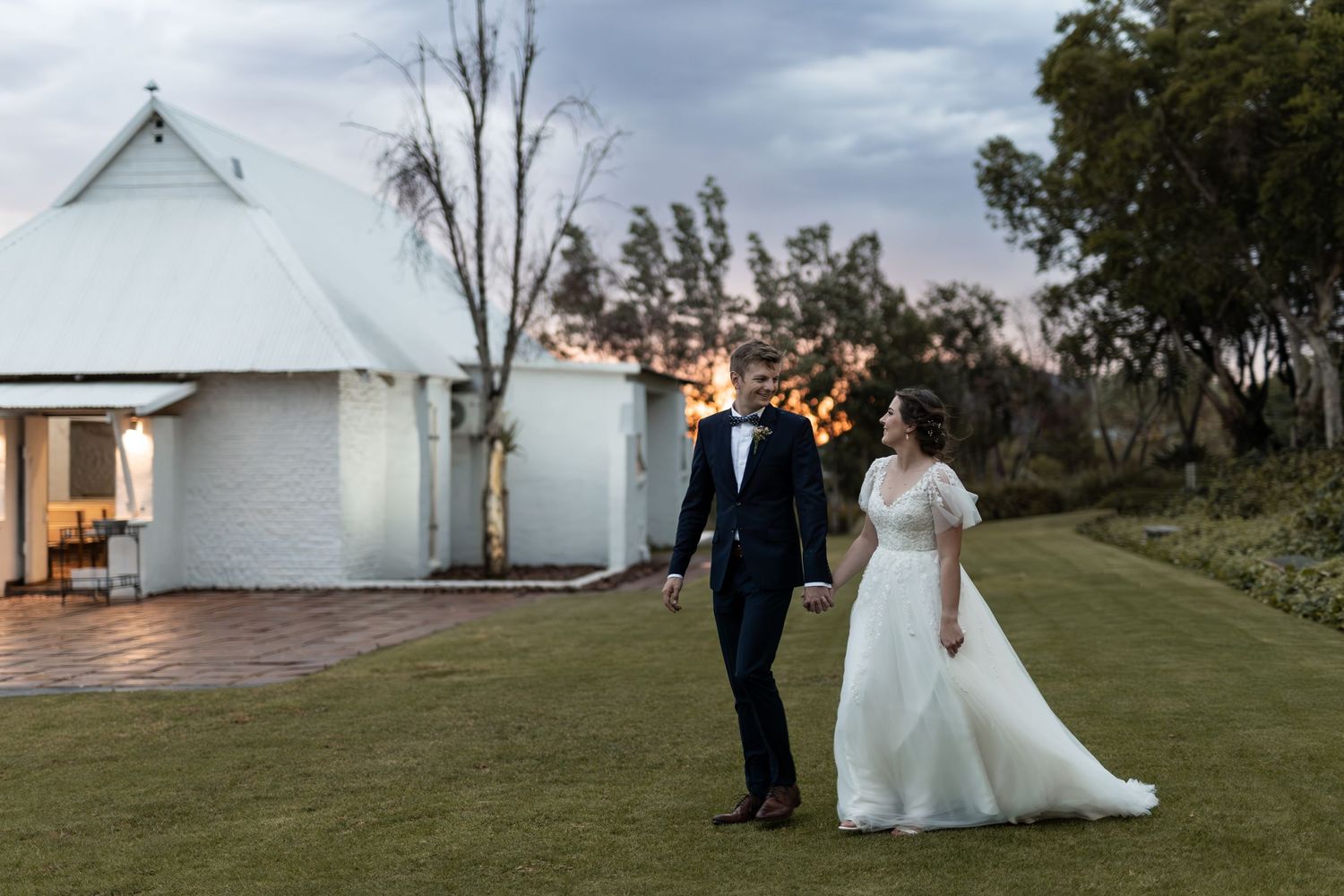 Wedding couple walking outside a white chapel venue at sunset with dramatic clouds overhead.
