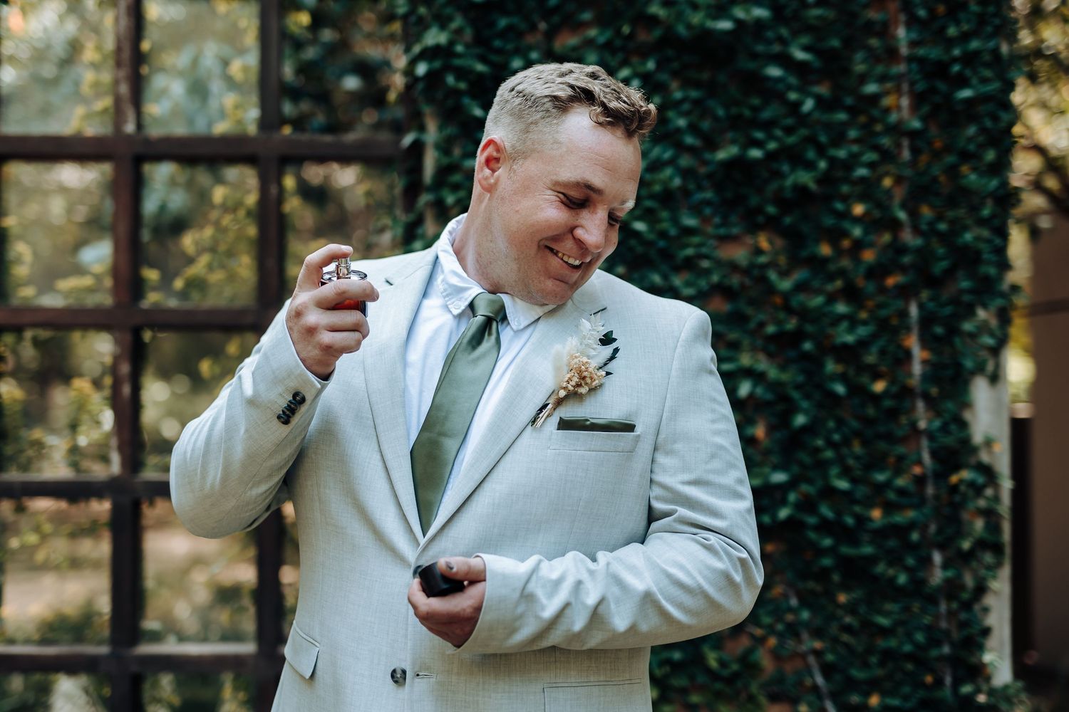 Person in light gray suit adjusts boutonniere while standing against garden backdrop.