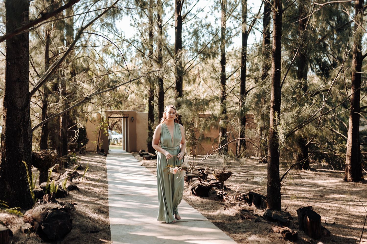 A wedding attendant in a sage green dress walks along a sunlit forest path surrounded by tall trees.