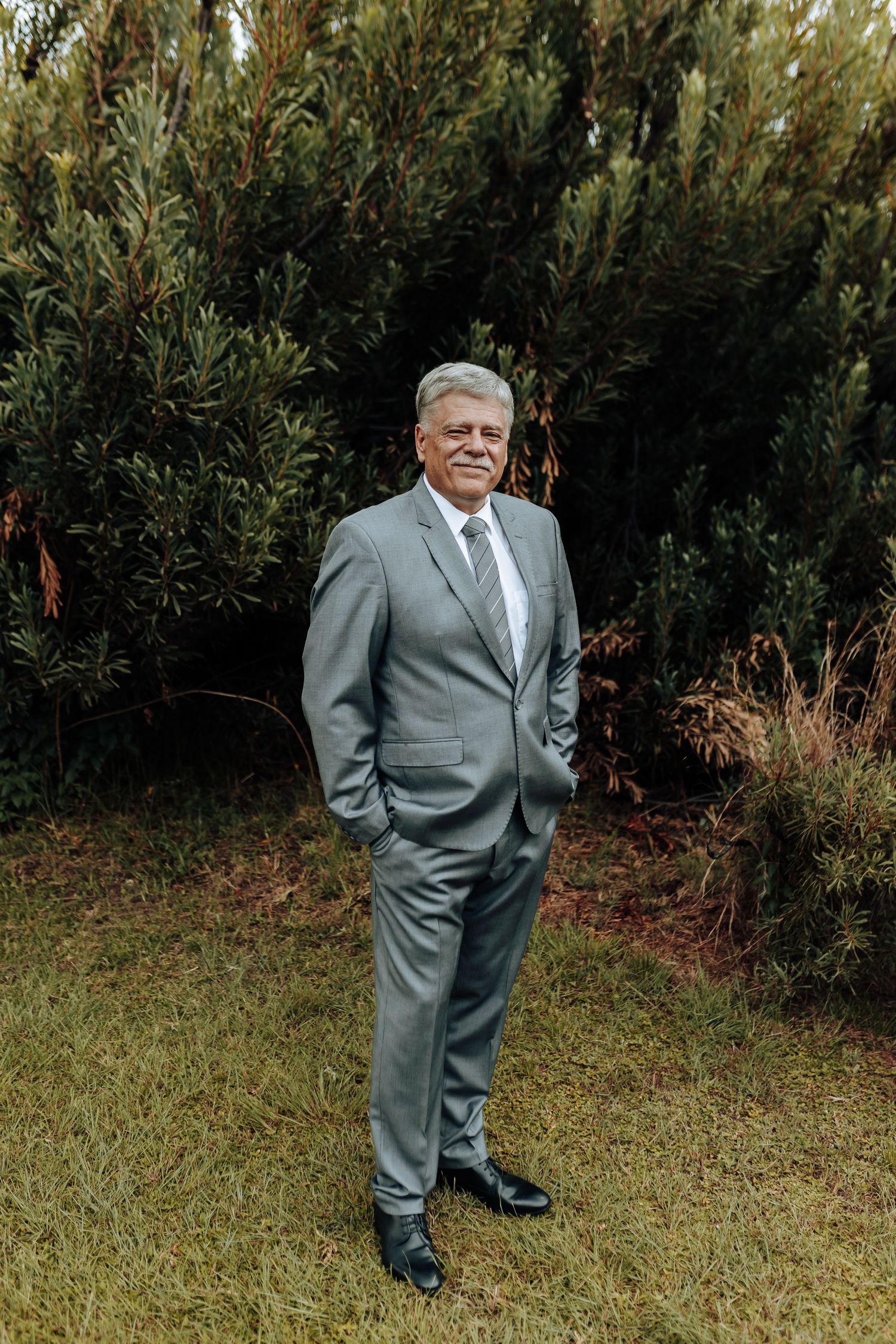 A person in a gray suit stands confidently in front of an evergreen hedge.