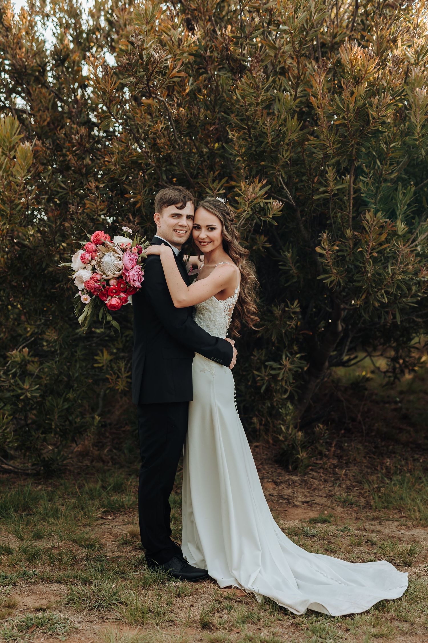Romantic wedding portrait of couple embracing against greenery with pink bouquet.