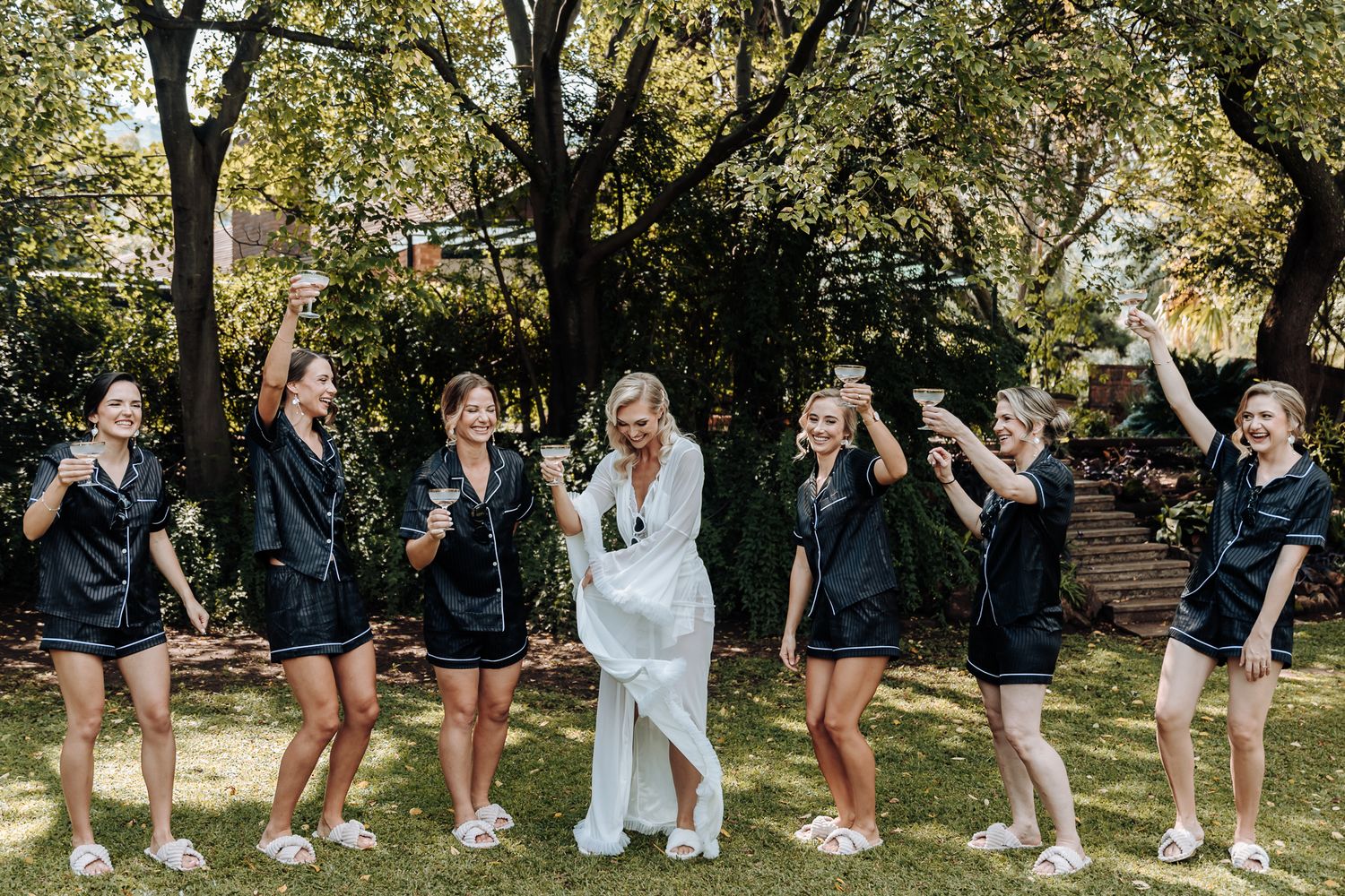 Bridesmaids in matching navy dresses and sunglasses celebrate with bride in white outdoors.