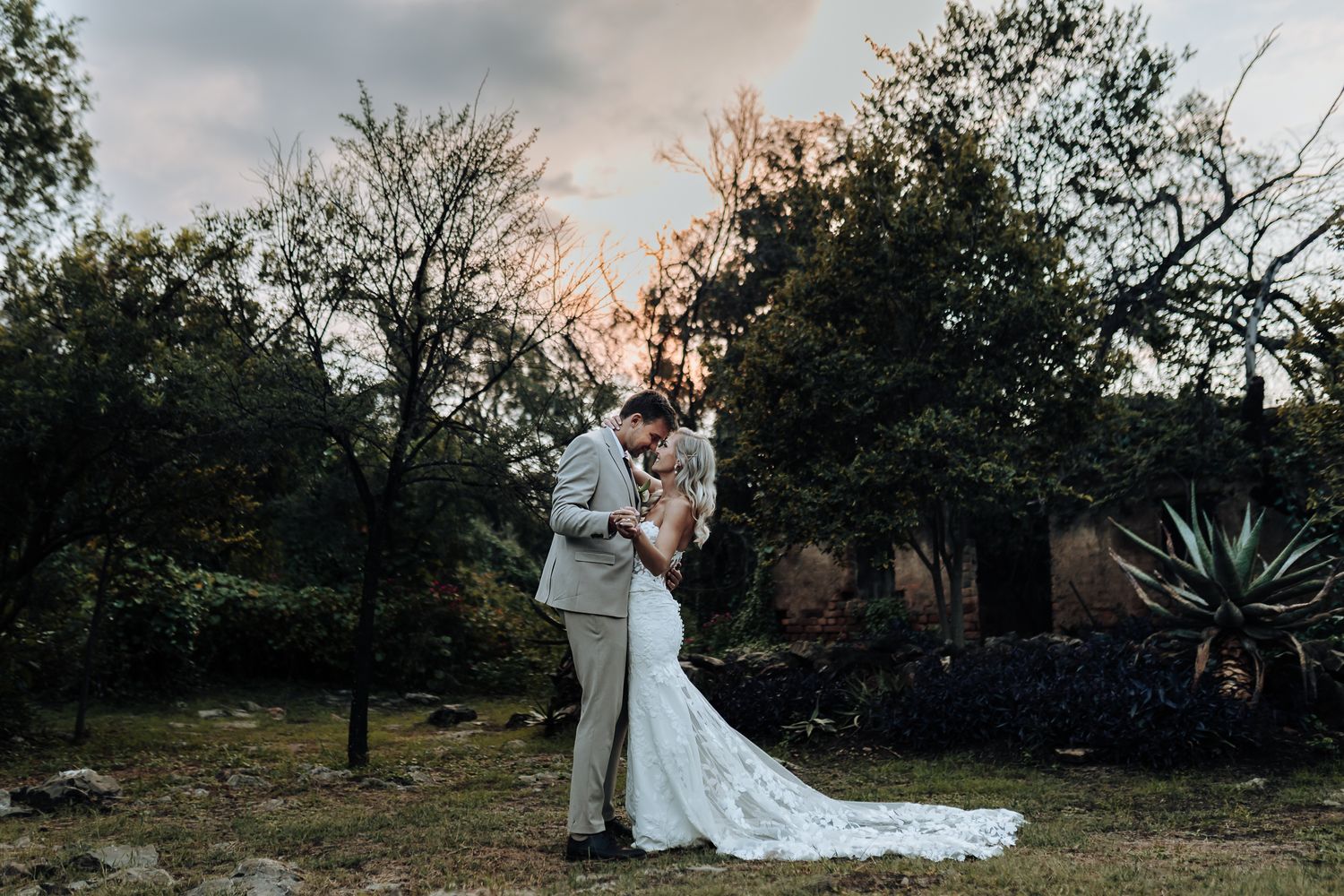 Silhouetted wedding couple embracing against sunset sky in garden setting.