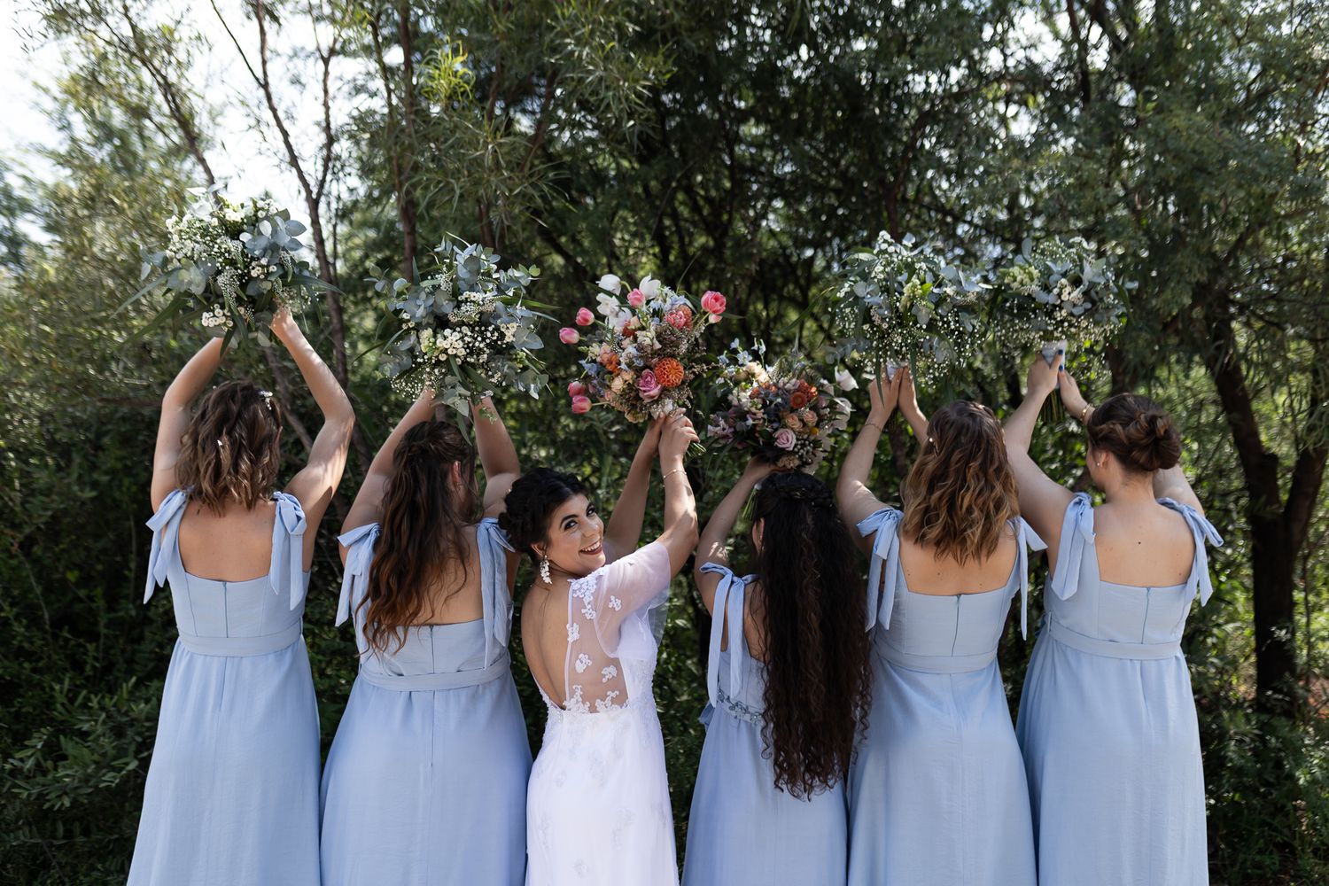 Wedding party raises their bouquets in celebration against a natural outdoor backdrop.
