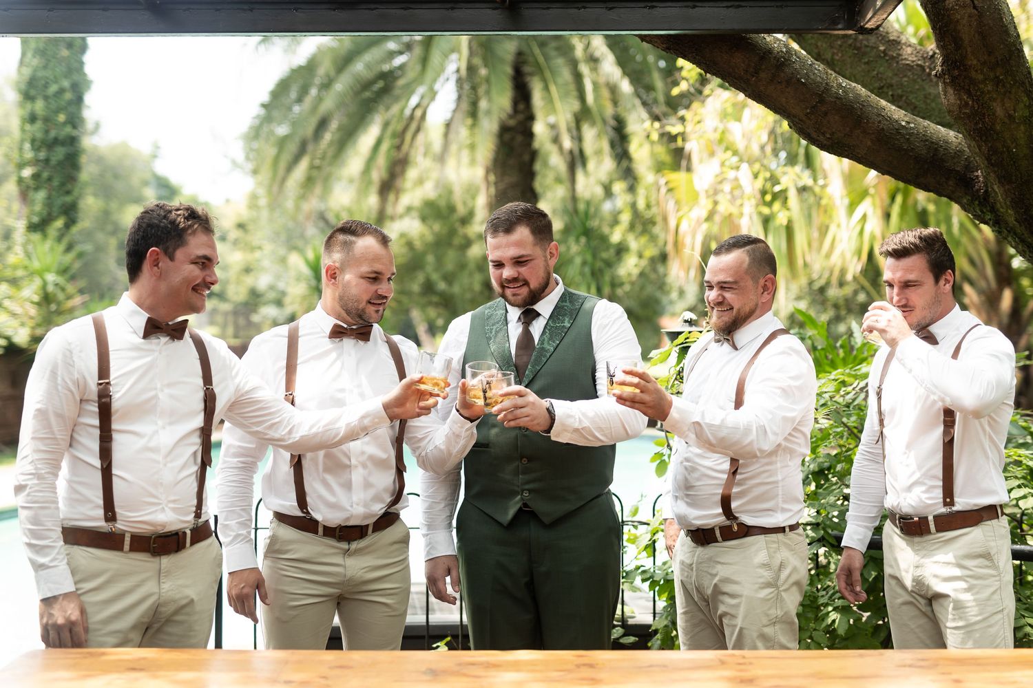 Groomsmen in suspenders toasting with drinks at an outdoor wedding celebration.