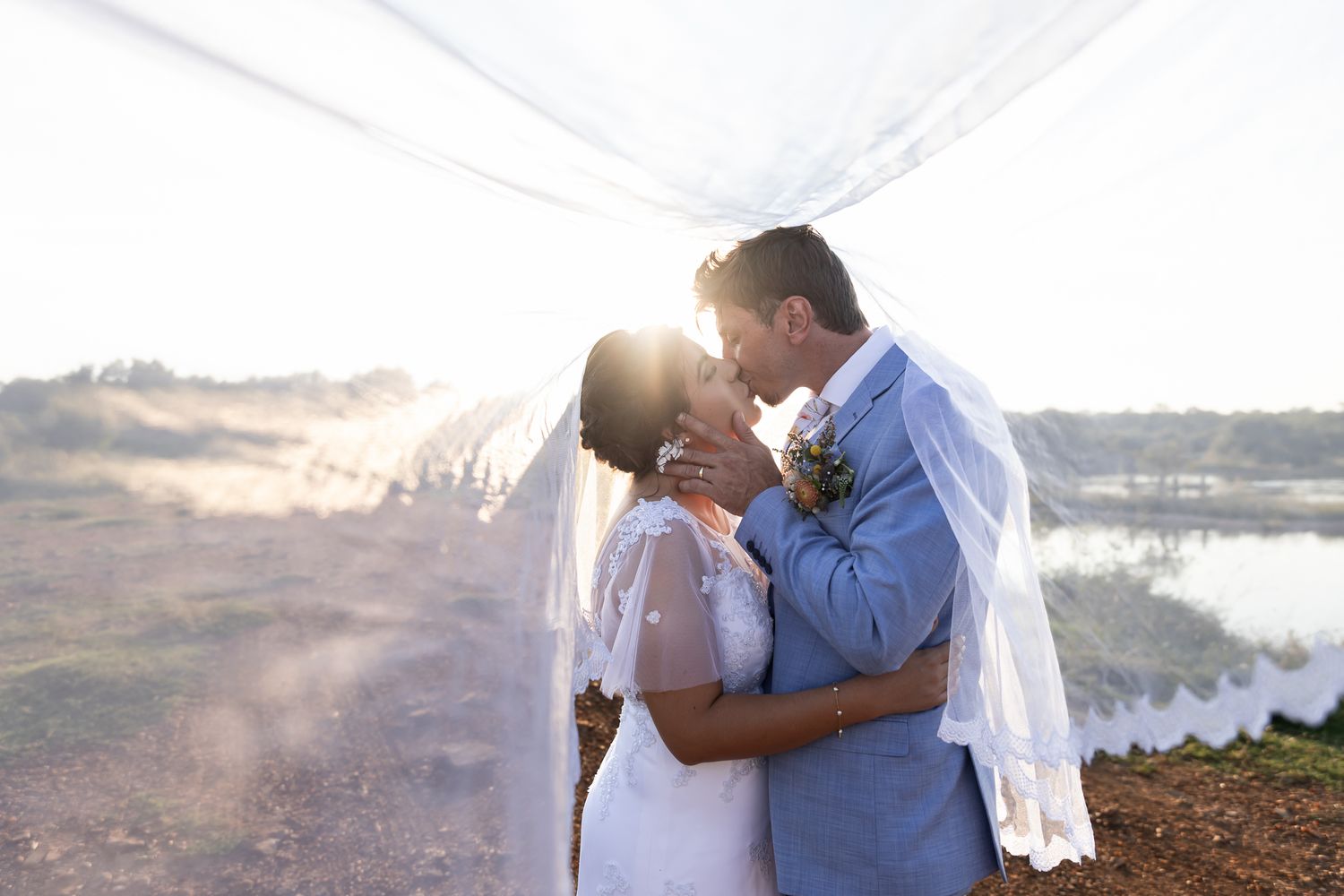 A couple shares a romantic moment under flowing white fabric near a lake at sunset.