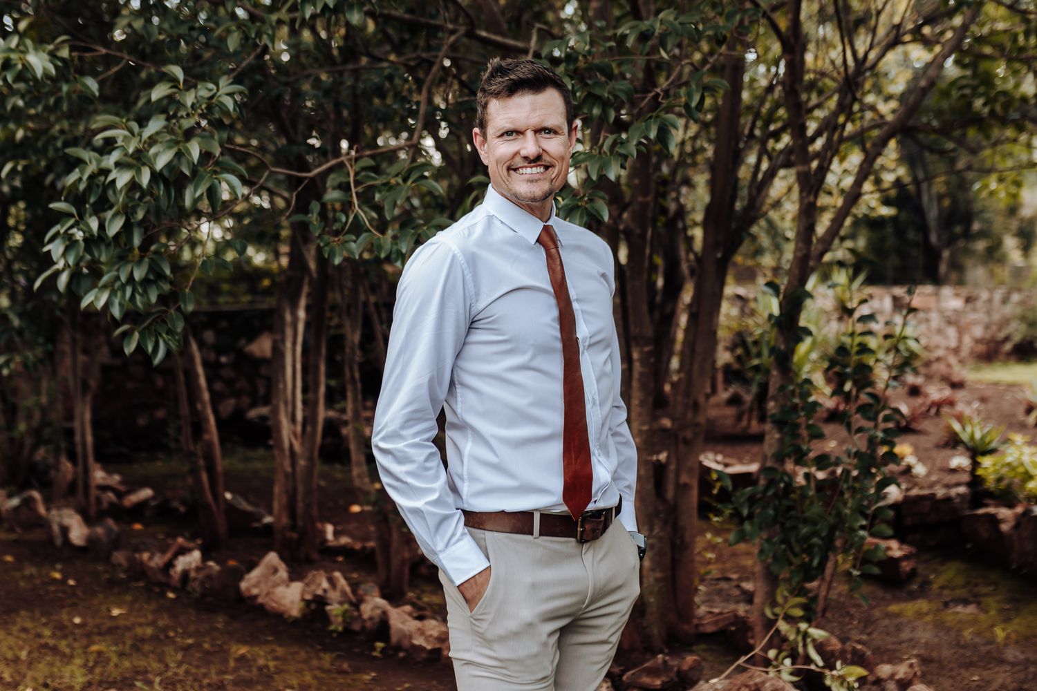 Man in light blue dress shirt and rust-colored tie poses confidently in outdoor woodland setting.