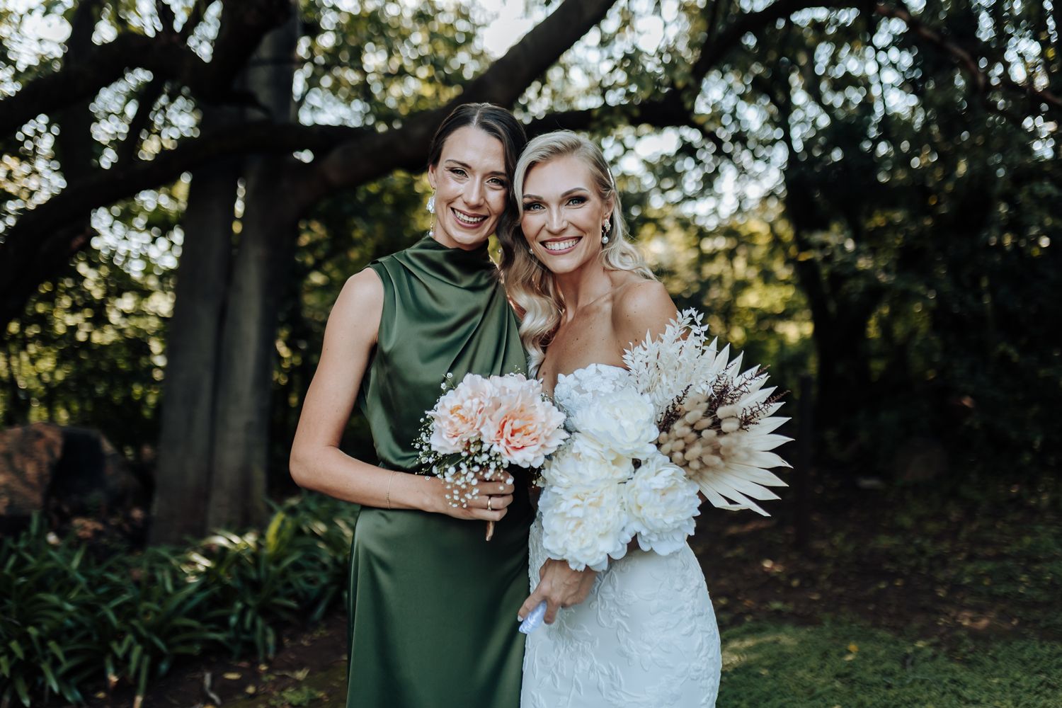 Two people pose with bouquets against a wooded backdrop, one in white lace and one in emerald green.