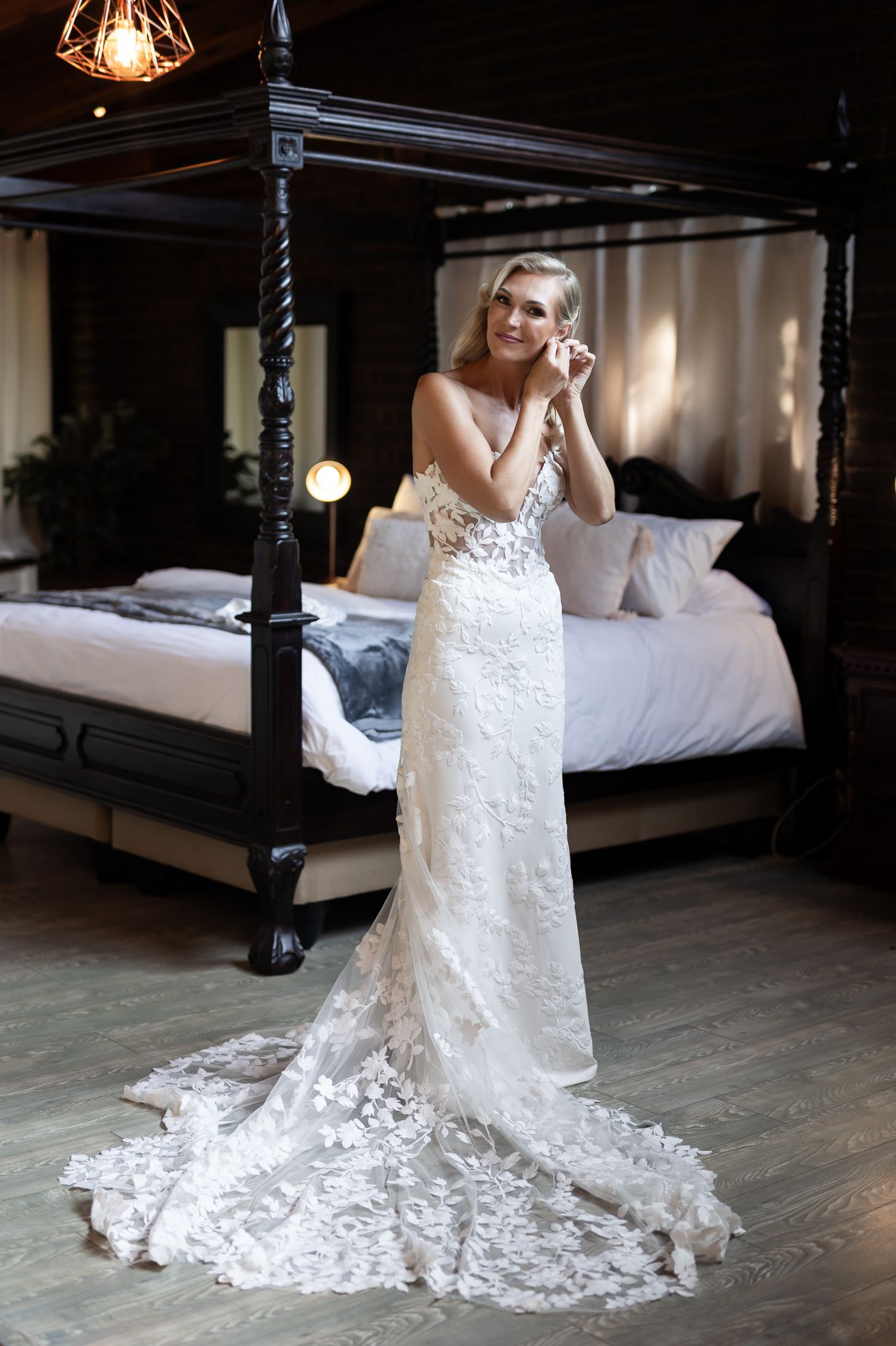 Bride in lace wedding gown with long train poses in rustic bedroom setting.