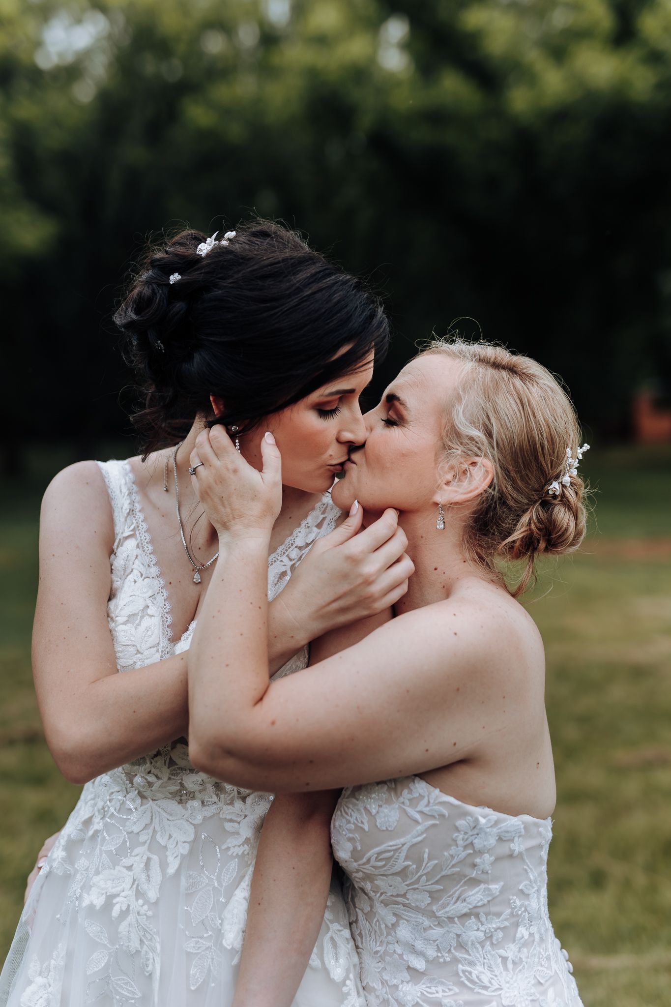 Two brides share an intimate moment in white lace wedding dresses against a natural outdoor backdrop.
