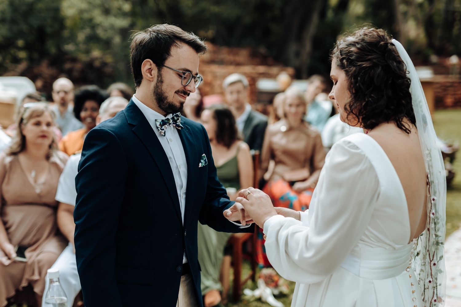 A close-up moment during an outdoor wedding ceremony with guests watching in the background.