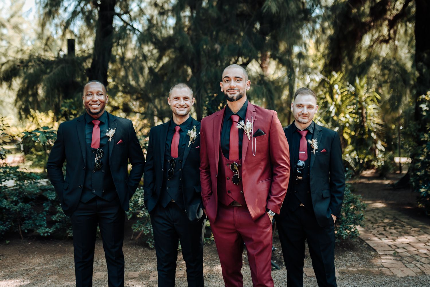 Groomsmen in black suits pose with groom in burgundy suit against evergreen trees.