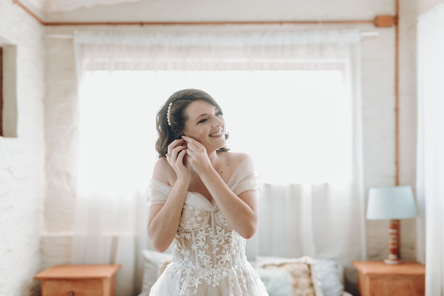 Elegant bride in a detailed lace wedding gown poses gracefully by the window.