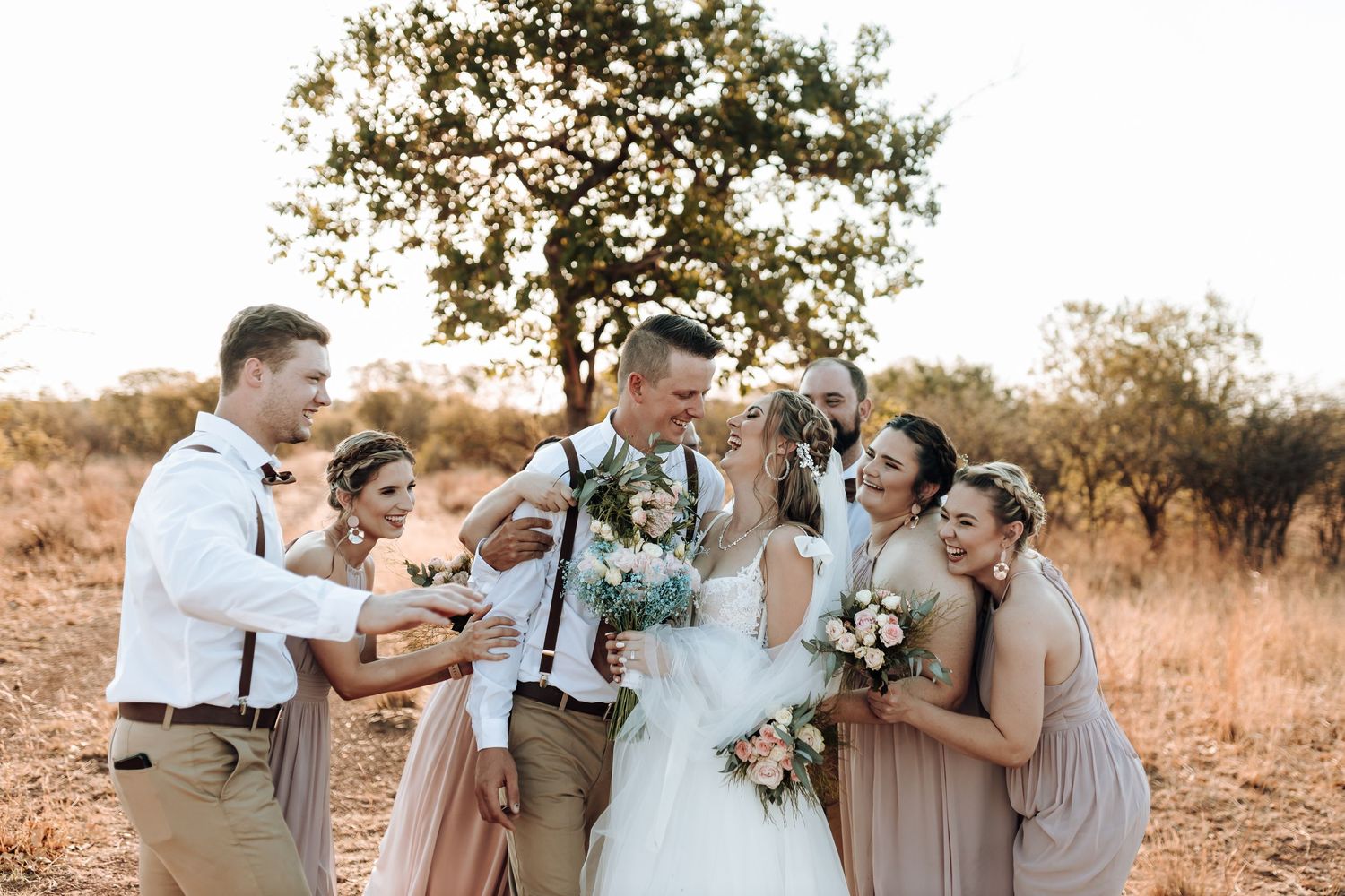 Wedding party shares candid laughing moment in outdoor setting.