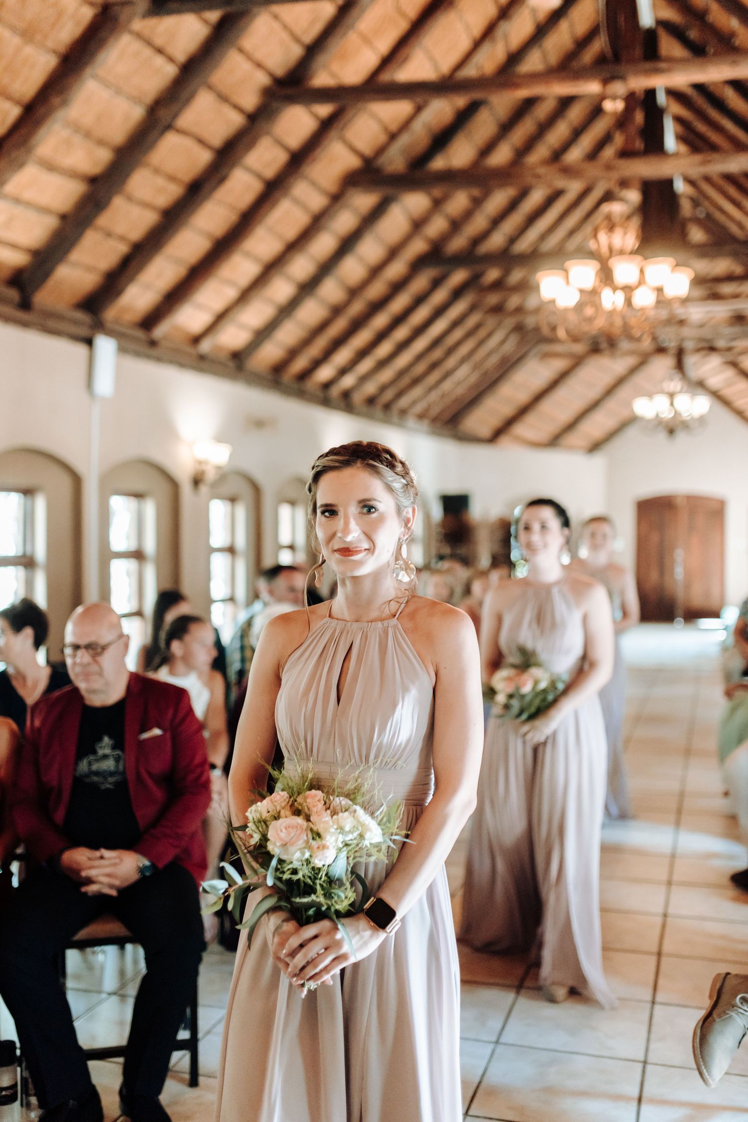 A bridesmaid in a flowing champagne dress holds a floral bouquet under wooden rafters at a wedding venue.