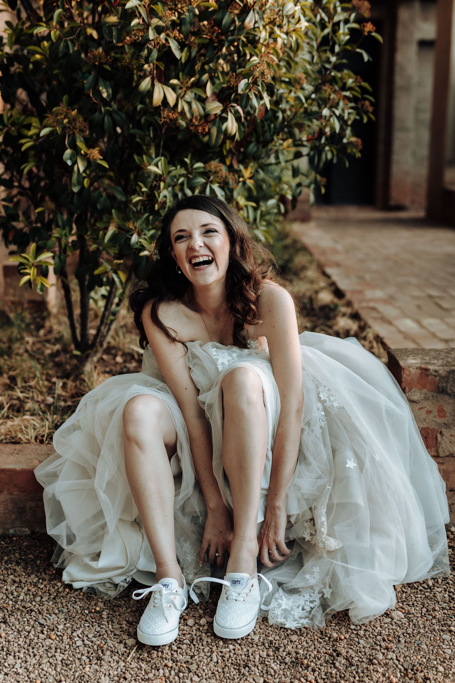 Bride wearing sneakers under wedding dress sits laughing on garden steps.