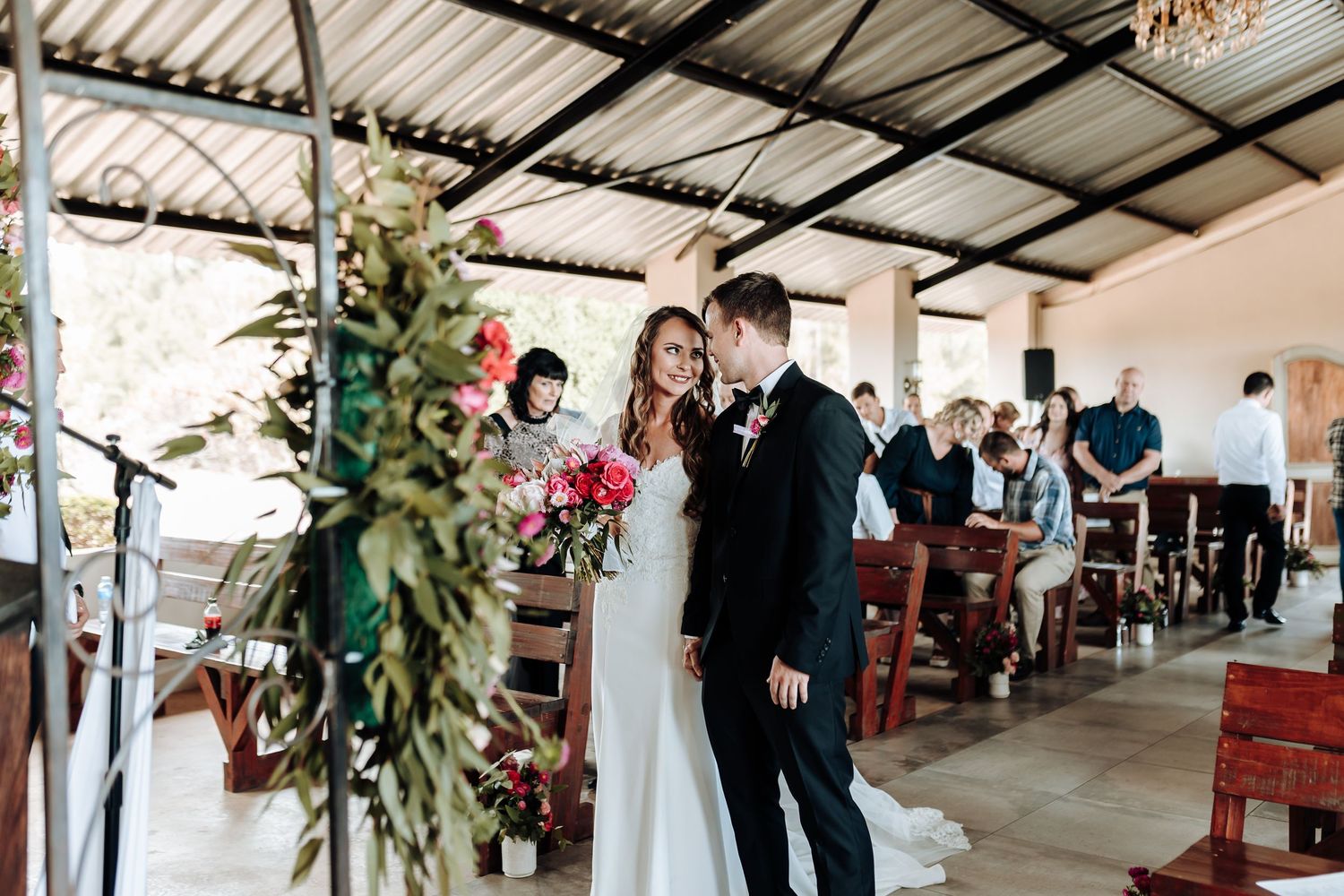 Wedding ceremony in progress with floral decorations and wooden pews in covered venue.