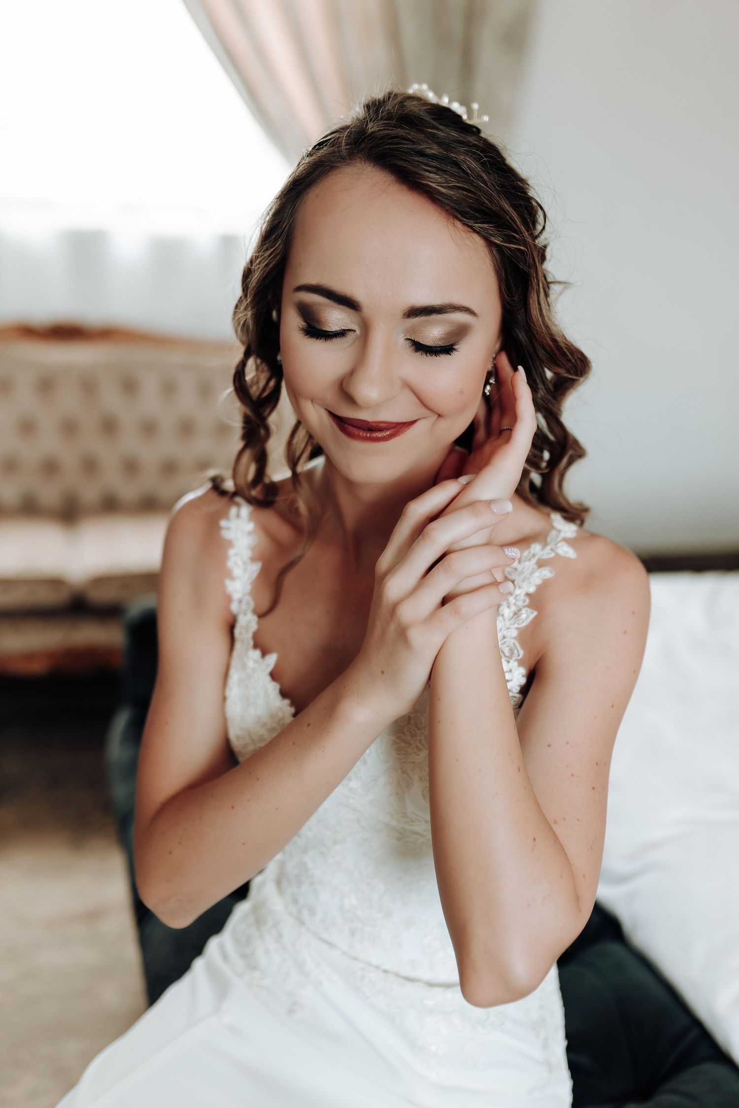 Bride in white lace dress with delicate jewelry touches her face with serene expression.
