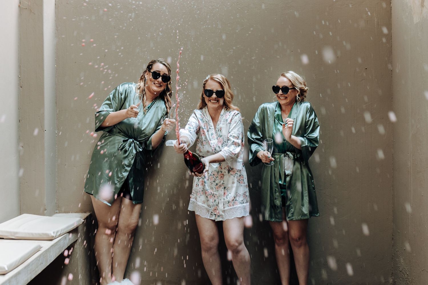 Friends in matching robes laugh as champagne sprays through the air during celebration.
