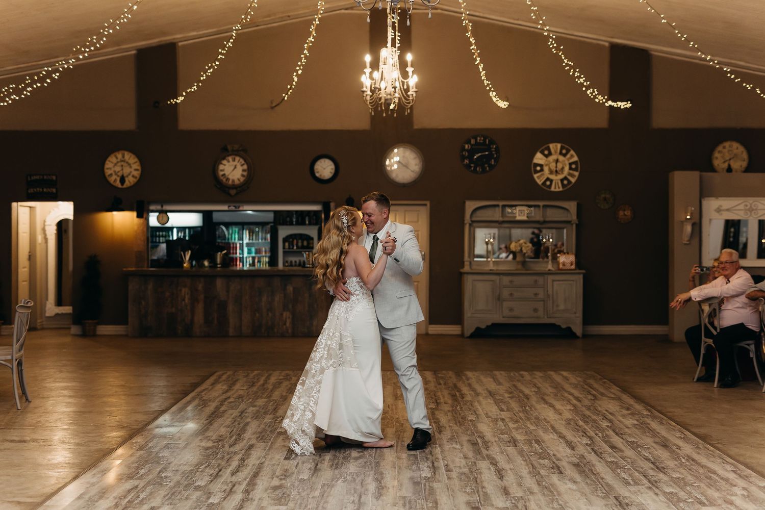 Couple dancing together in a warmly lit reception venue with wooden floors and fairy lights.