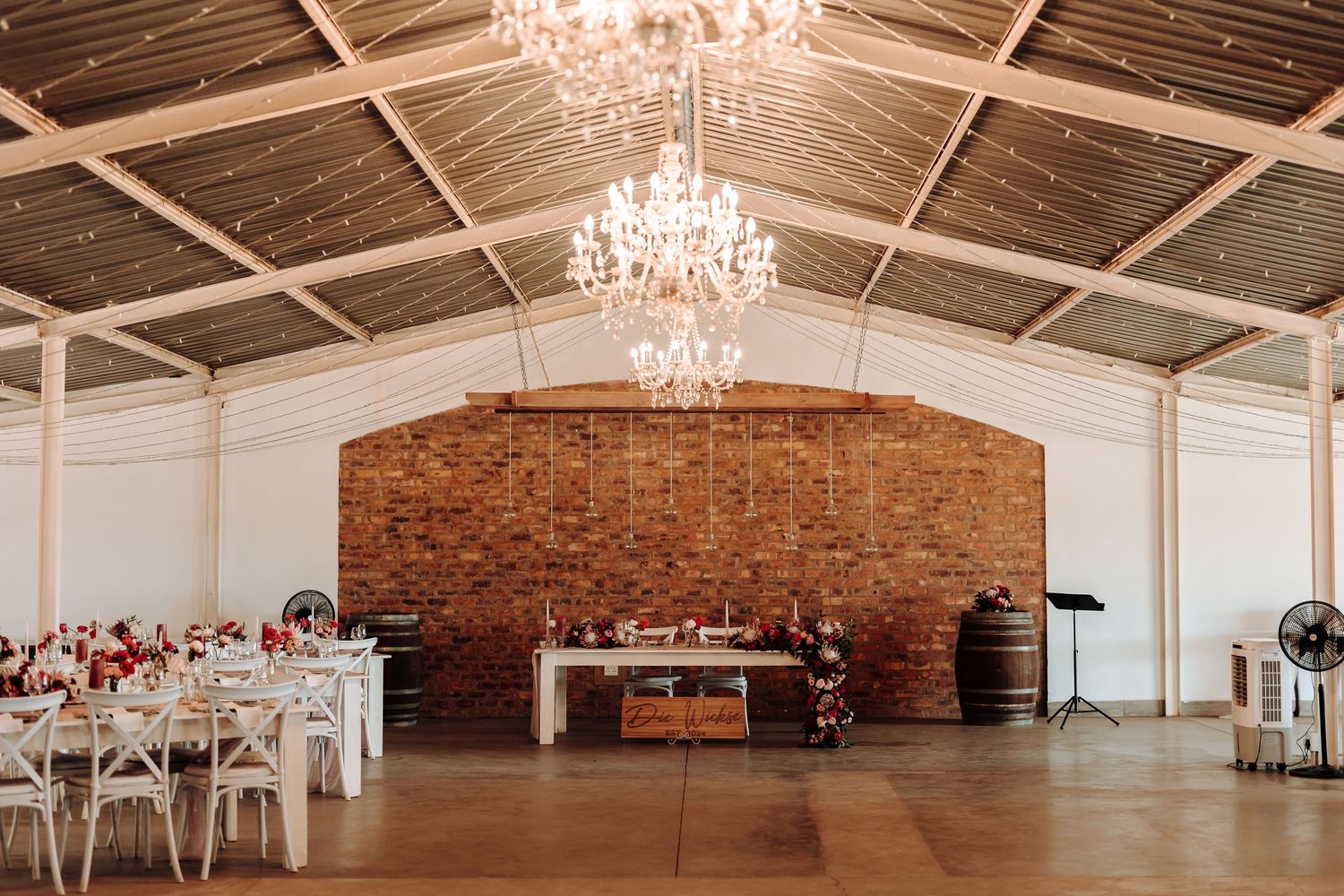 Elegant wedding reception hall with crystal chandeliers, exposed brick wall and white cross-back chairs.
