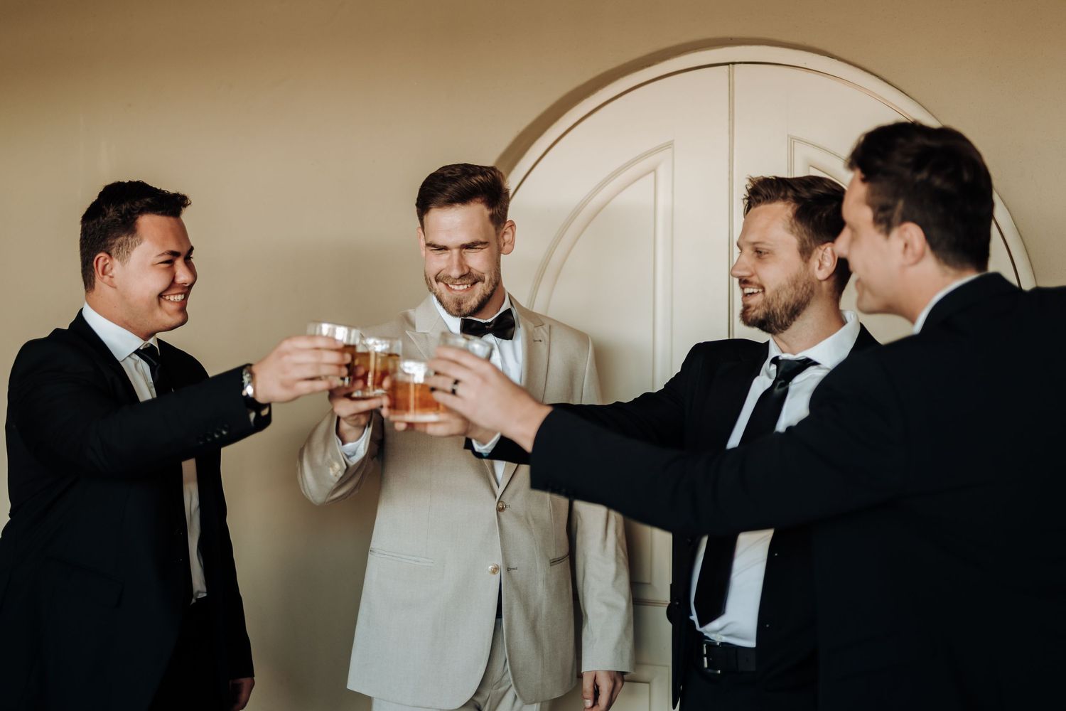 Groomsmen in formal attire toast with drinks during wedding preparation.