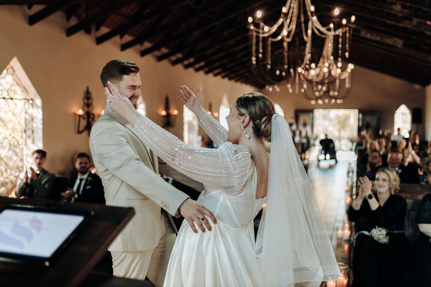 A joyful first dance moment between newlyweds in an elegant venue with chandeliers and arched windows.