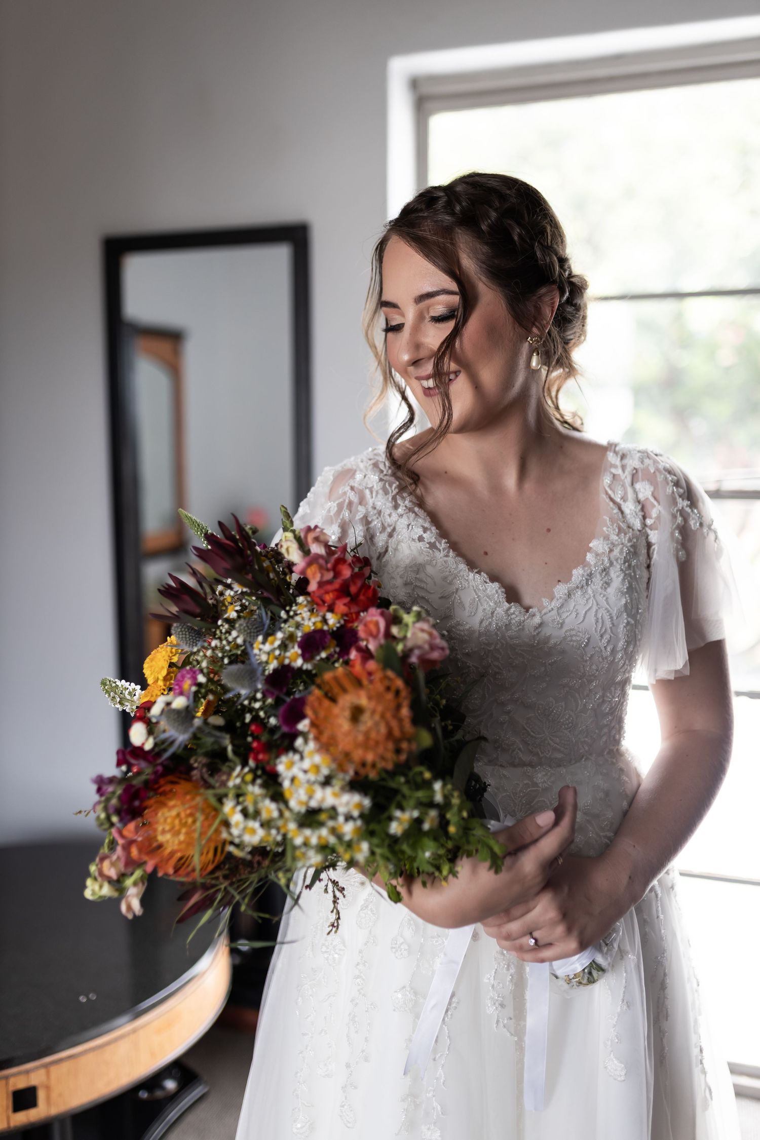 A bride wearing a delicate lace gown holds a colorful wildflower bouquet near a bright window.