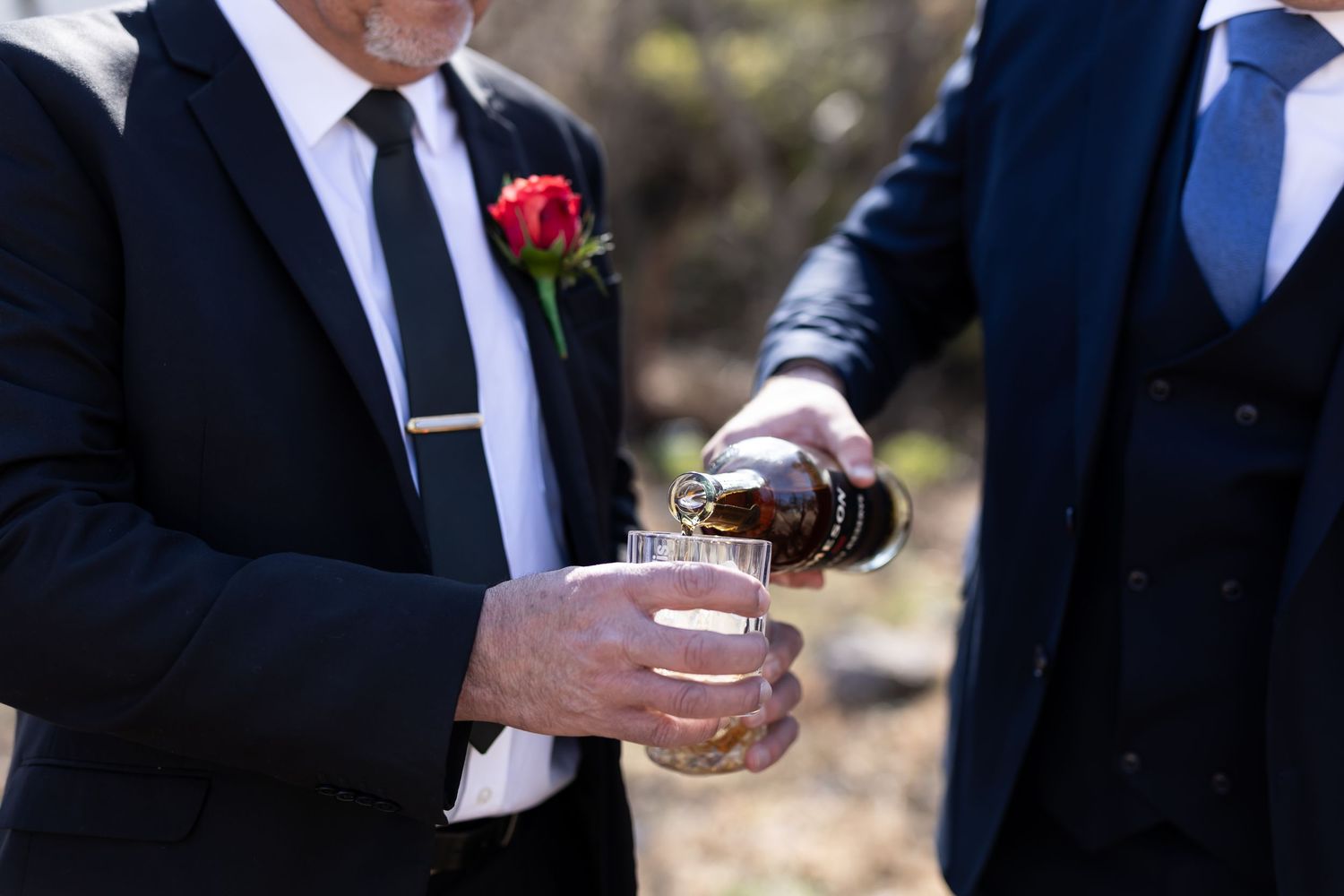 Two people in formal suits pour whiskey or bourbon into a small glass during an outdoor event.