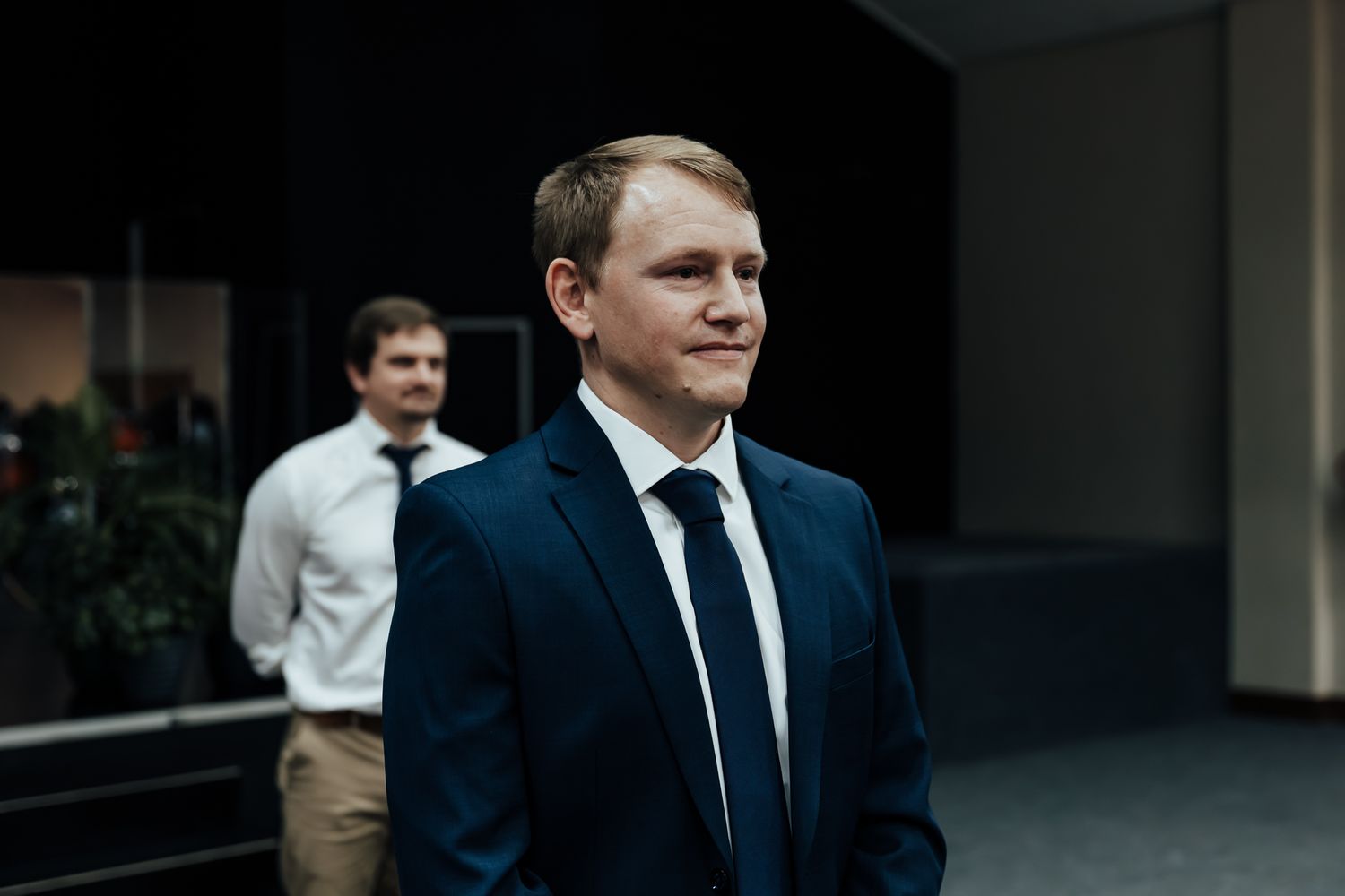 Dark-suited guests stand near a modern building entrance before a wedding ceremony.