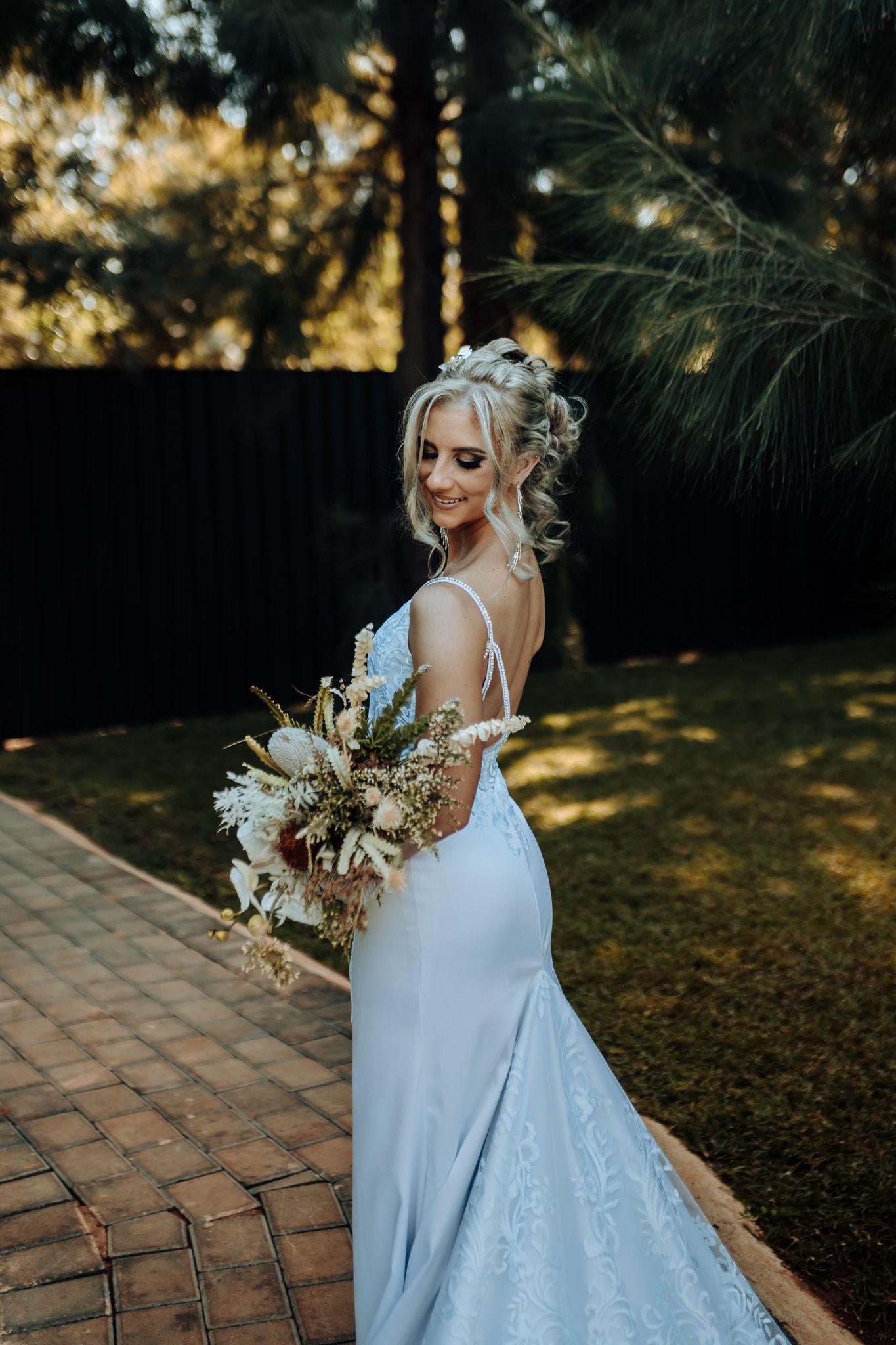 Bride with bouquet poses in fitted lace gown on garden path at sunset.