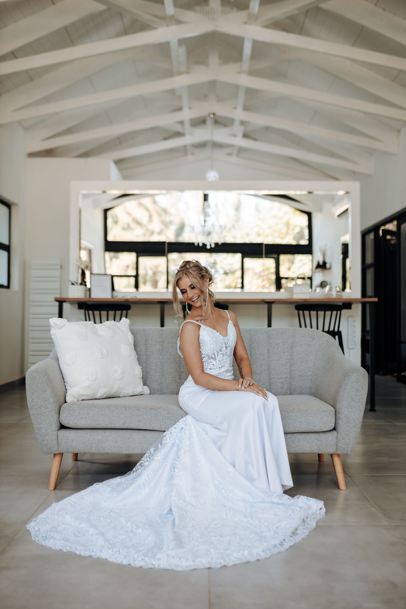 Elegant white lace wedding gown with fitted bodice and flowing train on gray sofa in modern bright venue.