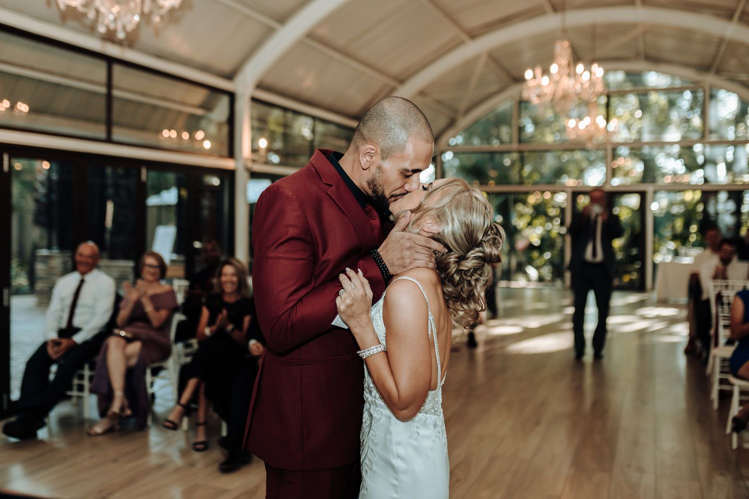 Romantic wedding moment captured in an elegant glass-enclosed venue with chandeliers.