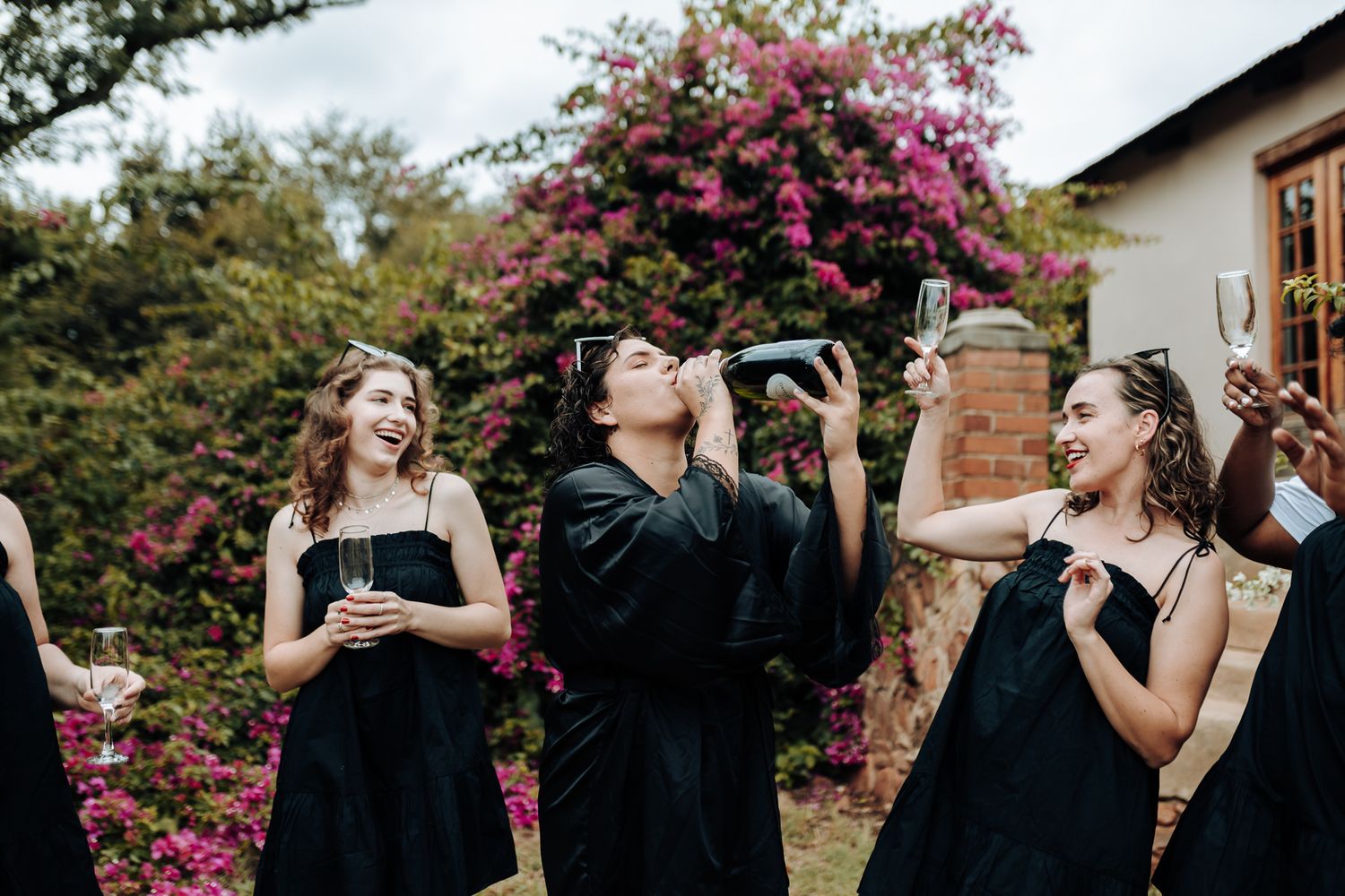 Friends in black dresses toasting and laughing in a flowering garden.