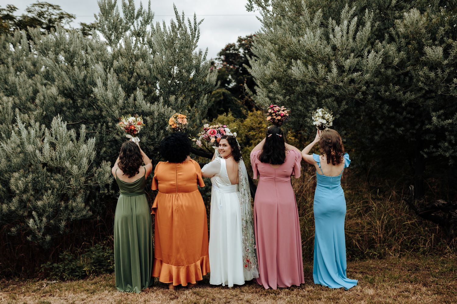 Back view of bridesmaids in different colored dresses standing in a row outdoors.