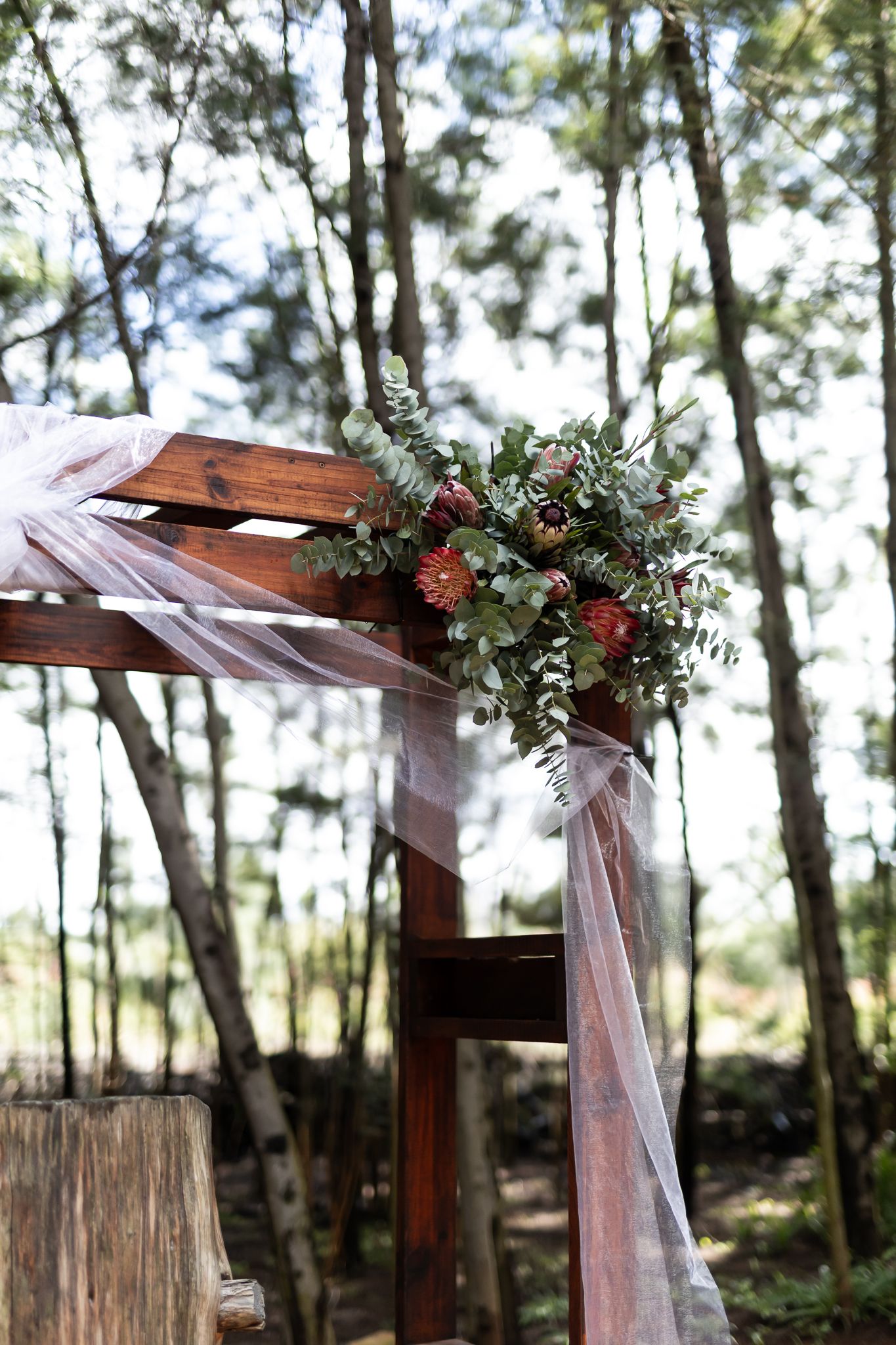 Wooden wedding arch corner decorated with red flowers, greenery and white tulle in forest setting.