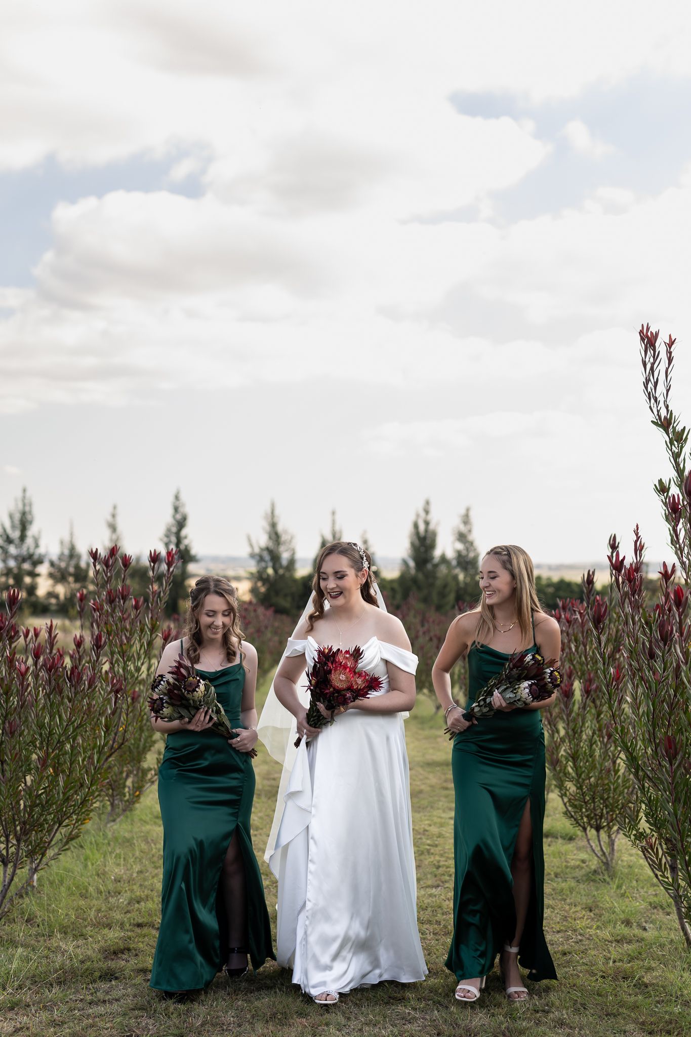 Three women in emerald green and white dresses pose with bouquets among flowering bushes.