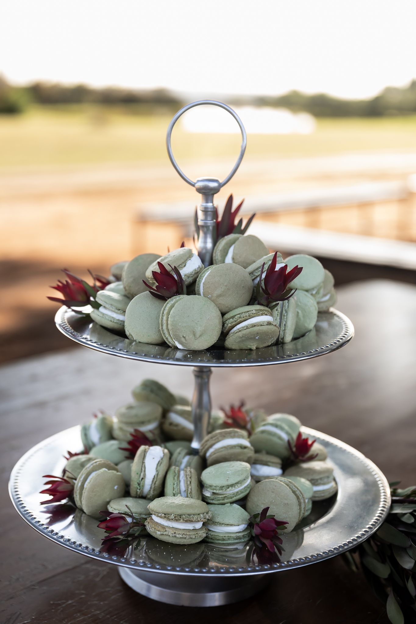 Two-tiered silver cake stand filled with mint-colored macarons and decorated with burgundy protea flowers.