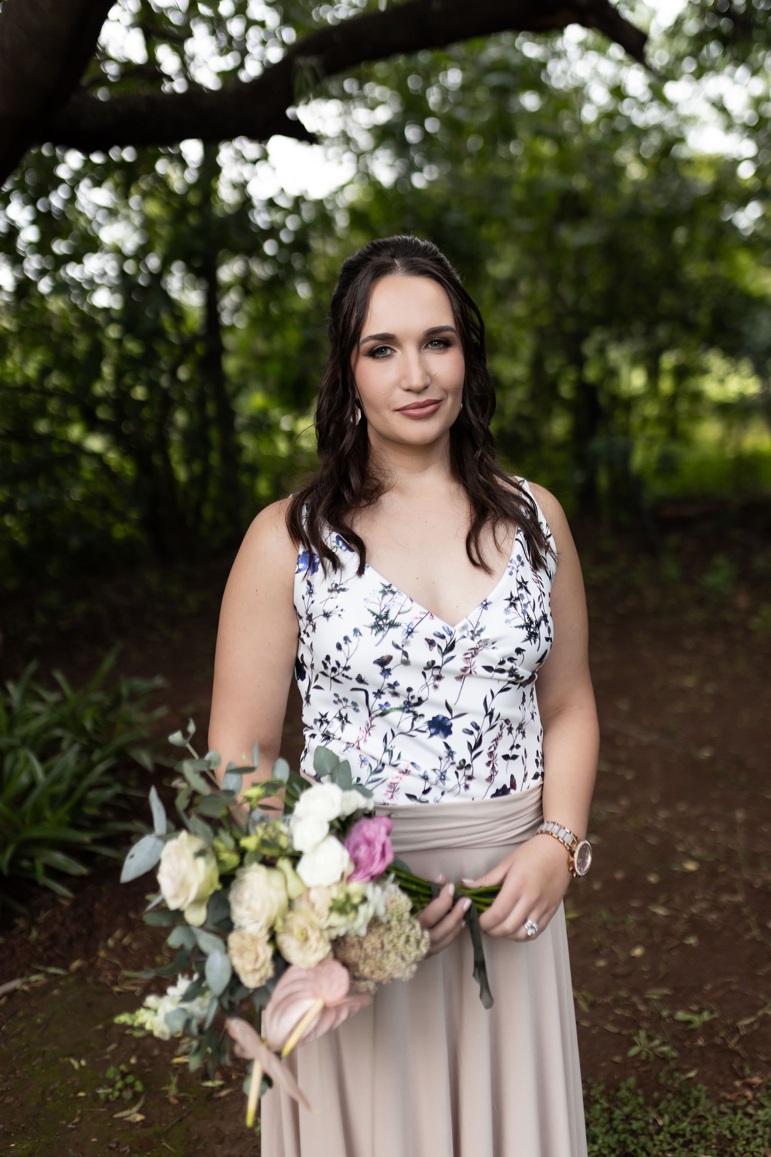 Bridesmaid wearing floral top and beige skirt poses with bouquet in outdoor garden setting.