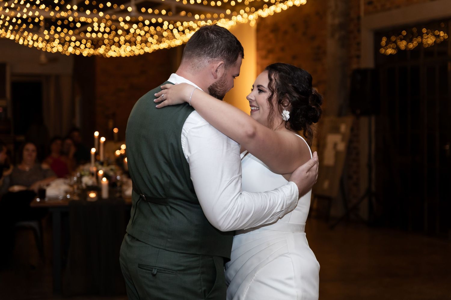 A tender first dance moment under warm twinkle lights in a rustic brick venue.