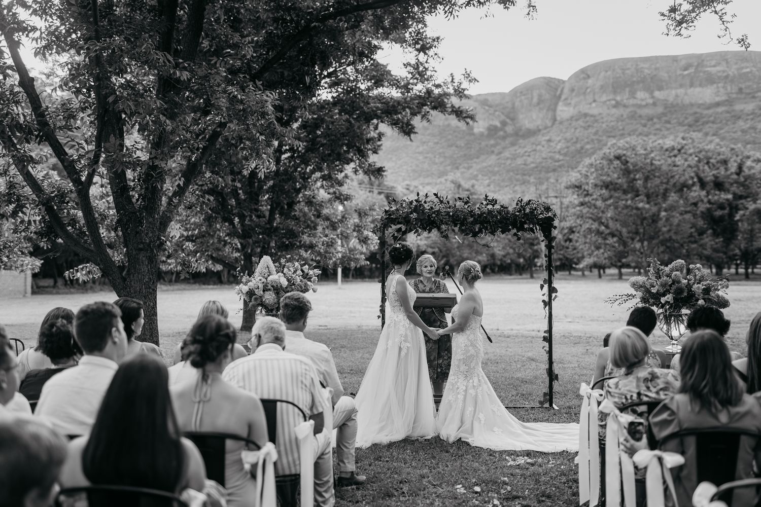 Black and white photo of outdoor wedding ceremony with mountain vista and guests seated under trees.